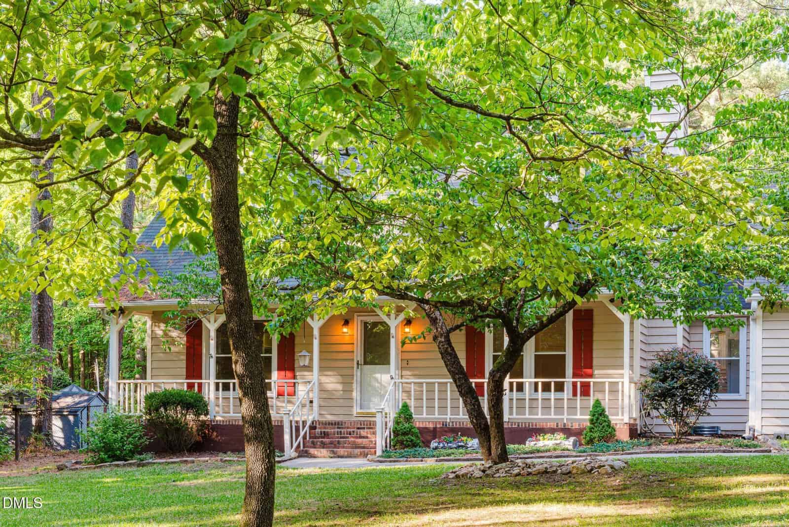 A charming white house with a wide front porch, red shutters, and steps leading to the entrance, surrounded by lush green trees and a well-kept lawn on a sunny day.