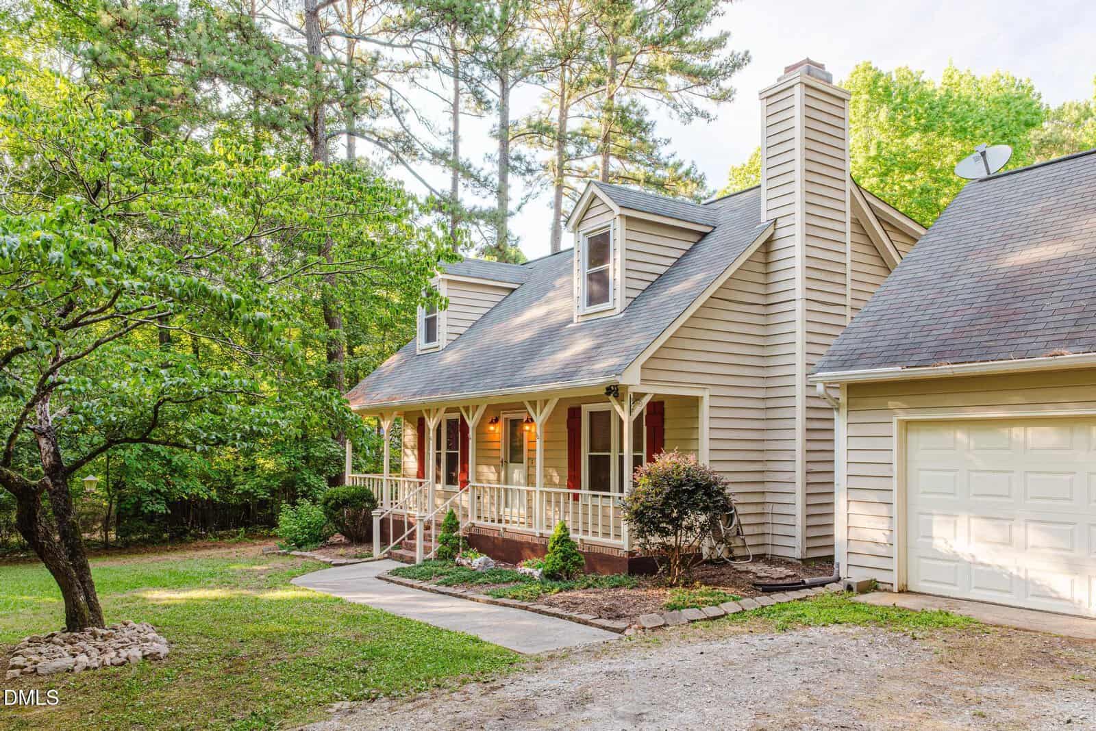 A beige, two-story house with a front porch, gabled roof, and red shutters is surrounded by trees and greenery. A paved walkway leads to the porch, and an attached garage is visible on the right.