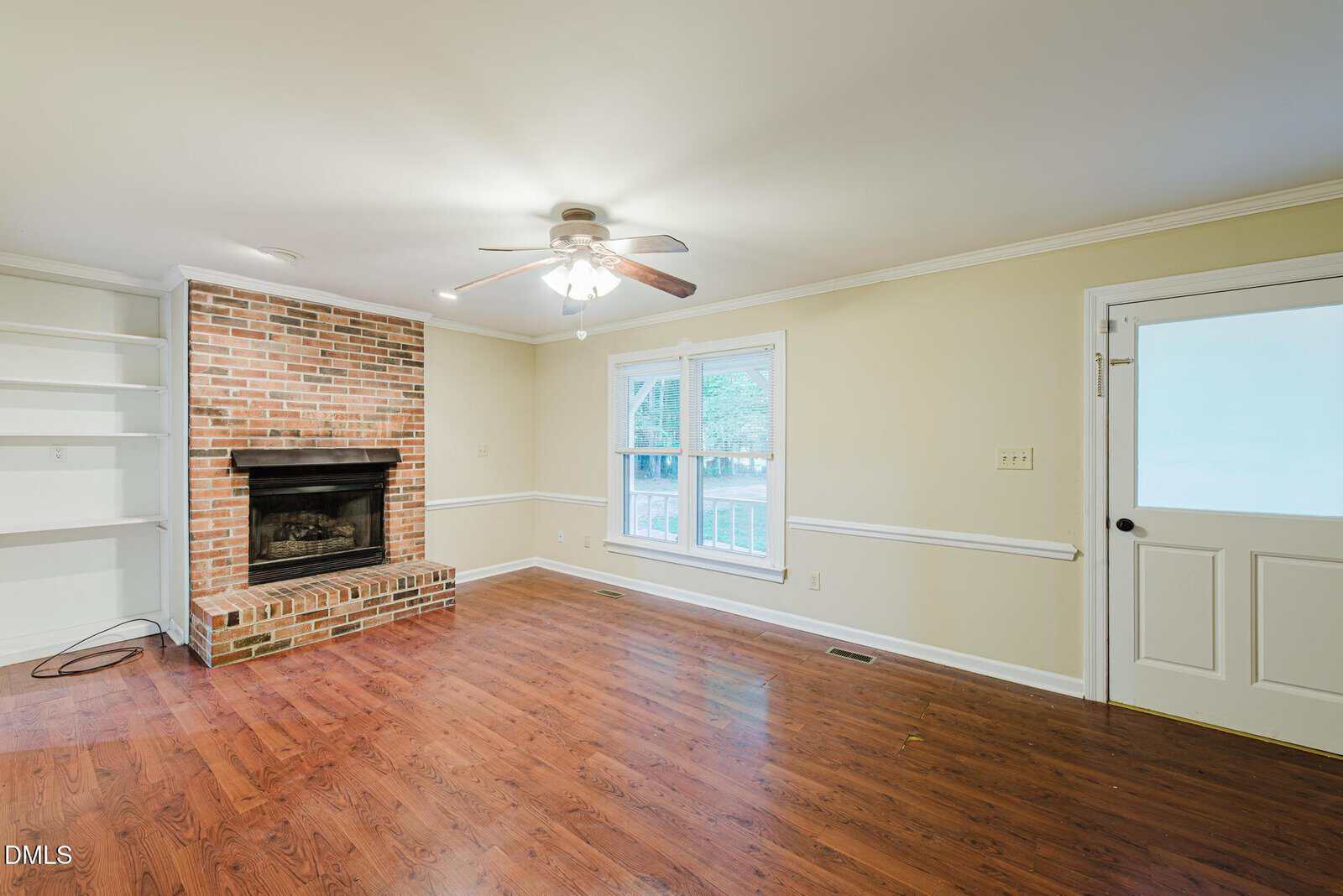 A bright living room with wood floors, a brick fireplace, built-in shelves, a ceiling fan, two large windows, and a white door with frosted glass.