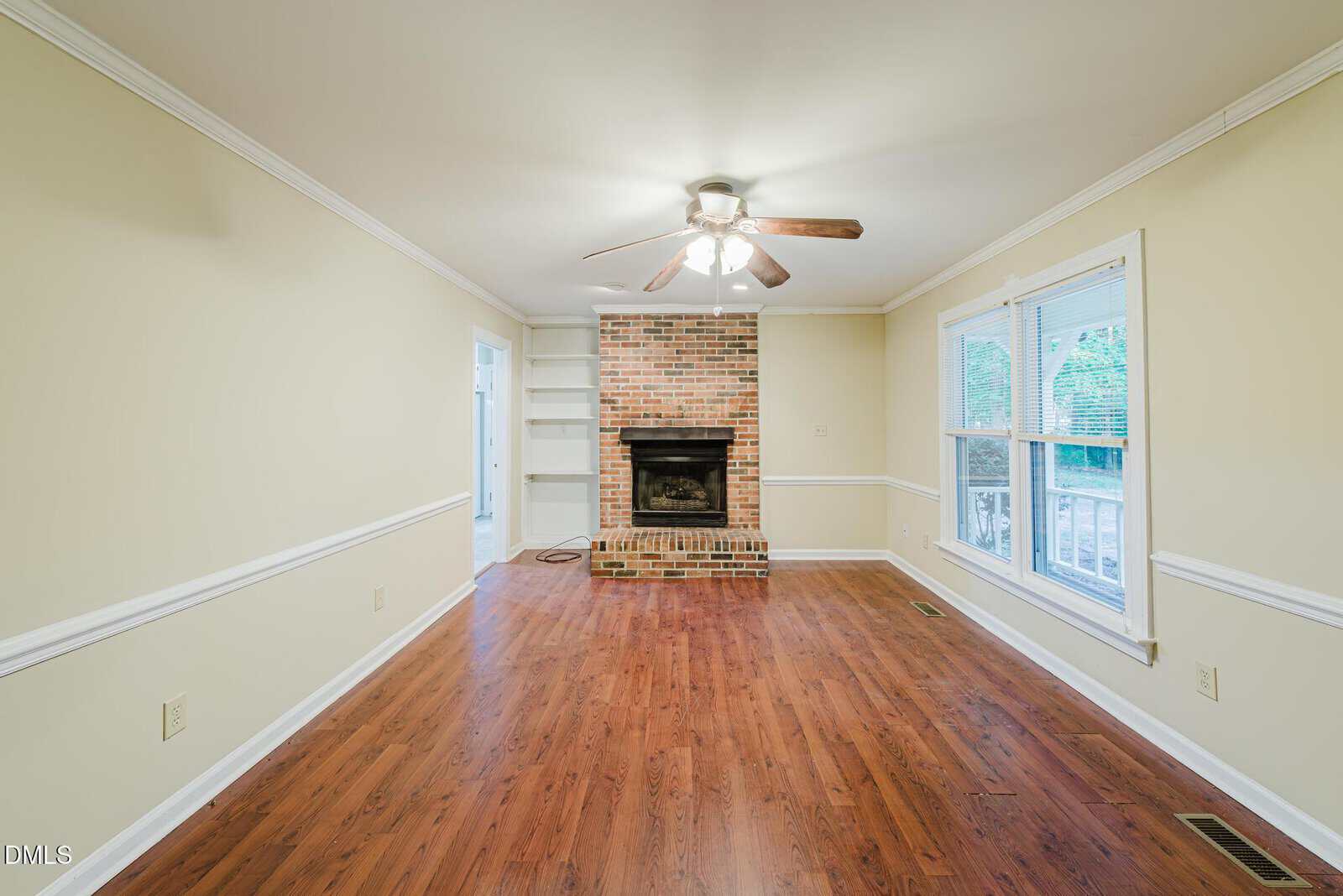A bright living room with hardwood floors, a brick fireplace, built-in shelves, a ceiling fan, and large windows letting in natural light.