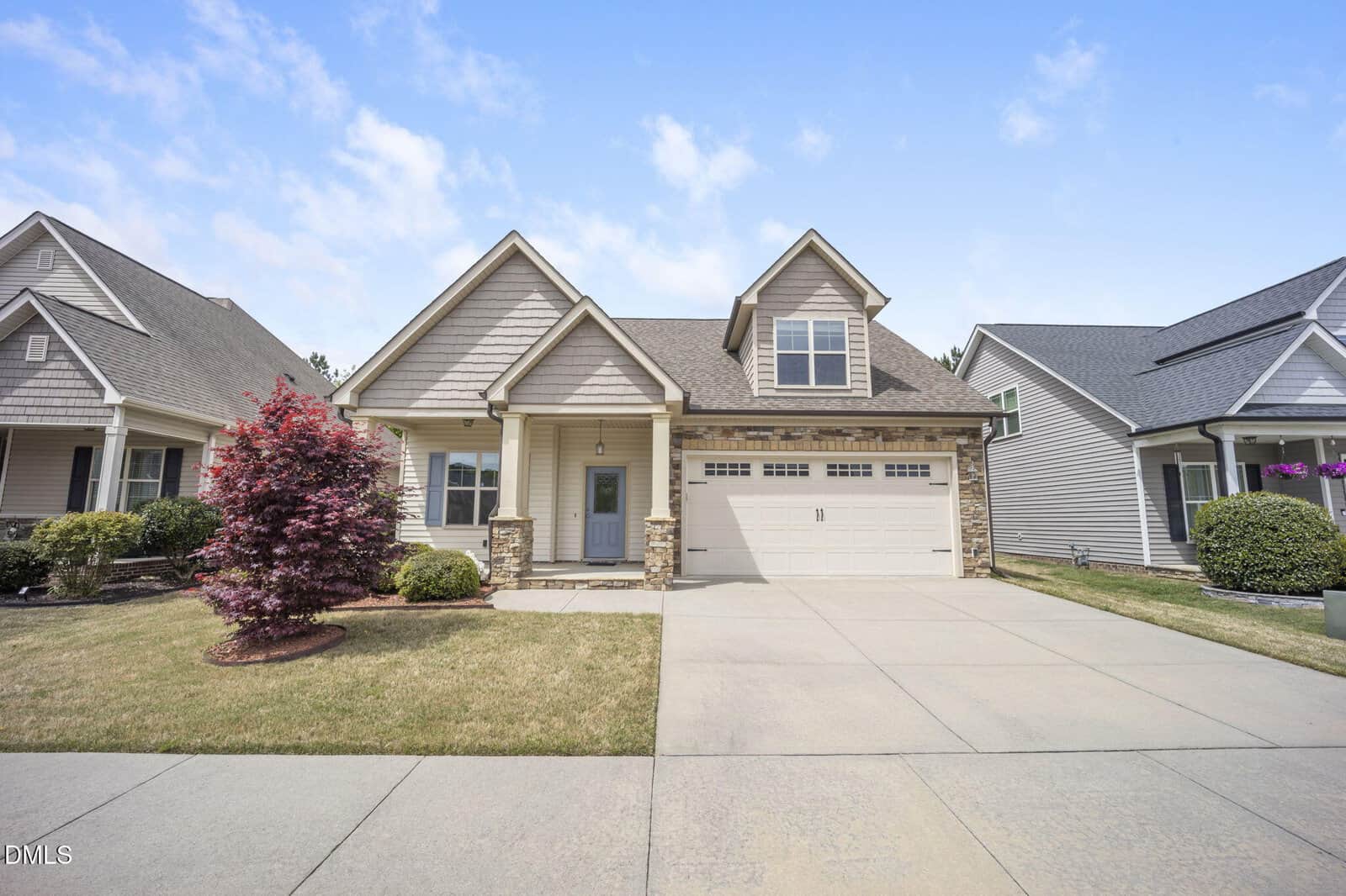 A modern suburban house at 100 Scarlet Bell Drive, Youngsville, features beige siding, stone accents, a covered porch, double garage, and a neatly maintained front yard with grass, bushes, and a red-leafed tree under a partly cloudy sky.