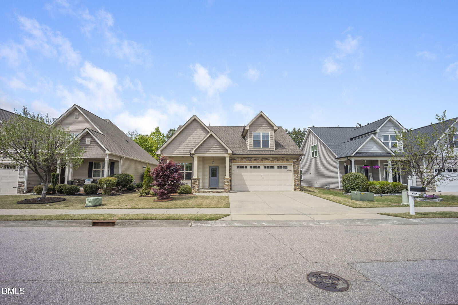 A suburban street with three modern single-family homes, each with a front lawn, driveway, and attached garage, under a blue sky with scattered clouds.