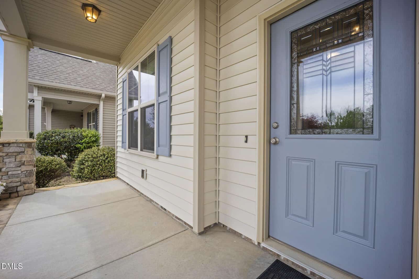 Covered entryway of a house with beige siding, a blue front door with glass panels, a small window, and a landscaped area with bushes to the left.