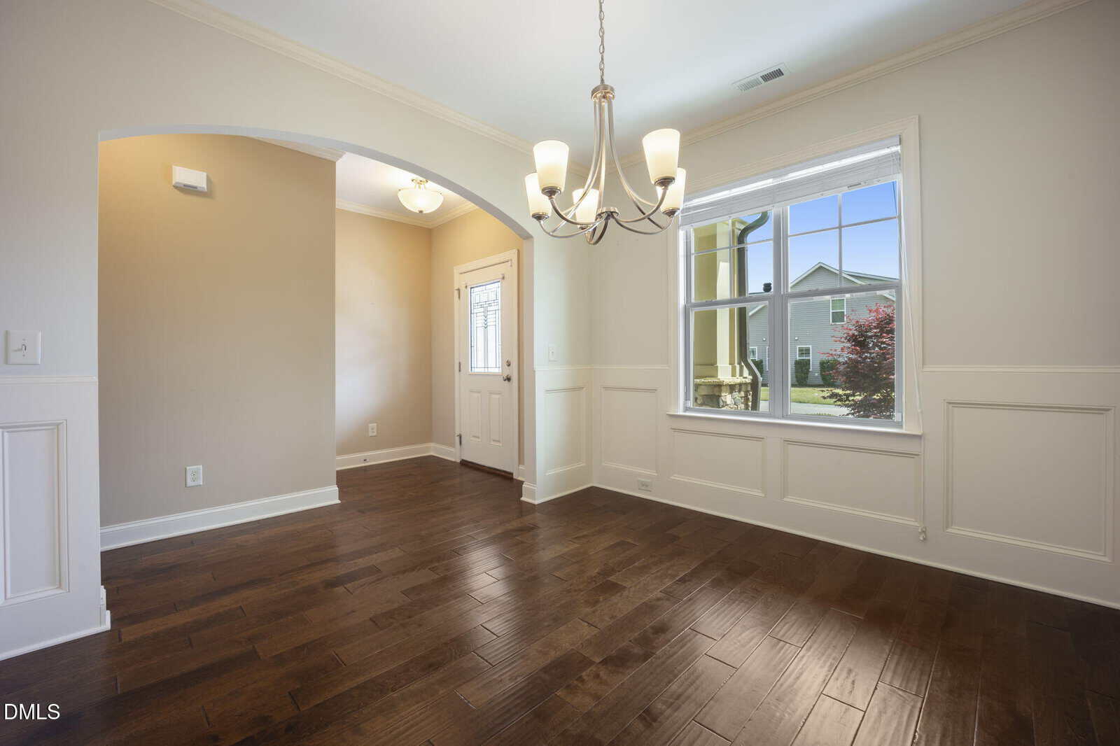 Bright dining room with hardwood floors, white wainscoting, a chandelier, and large window with a view of a green house and red bush. An arched doorway leads to the entryway with a white front door.
