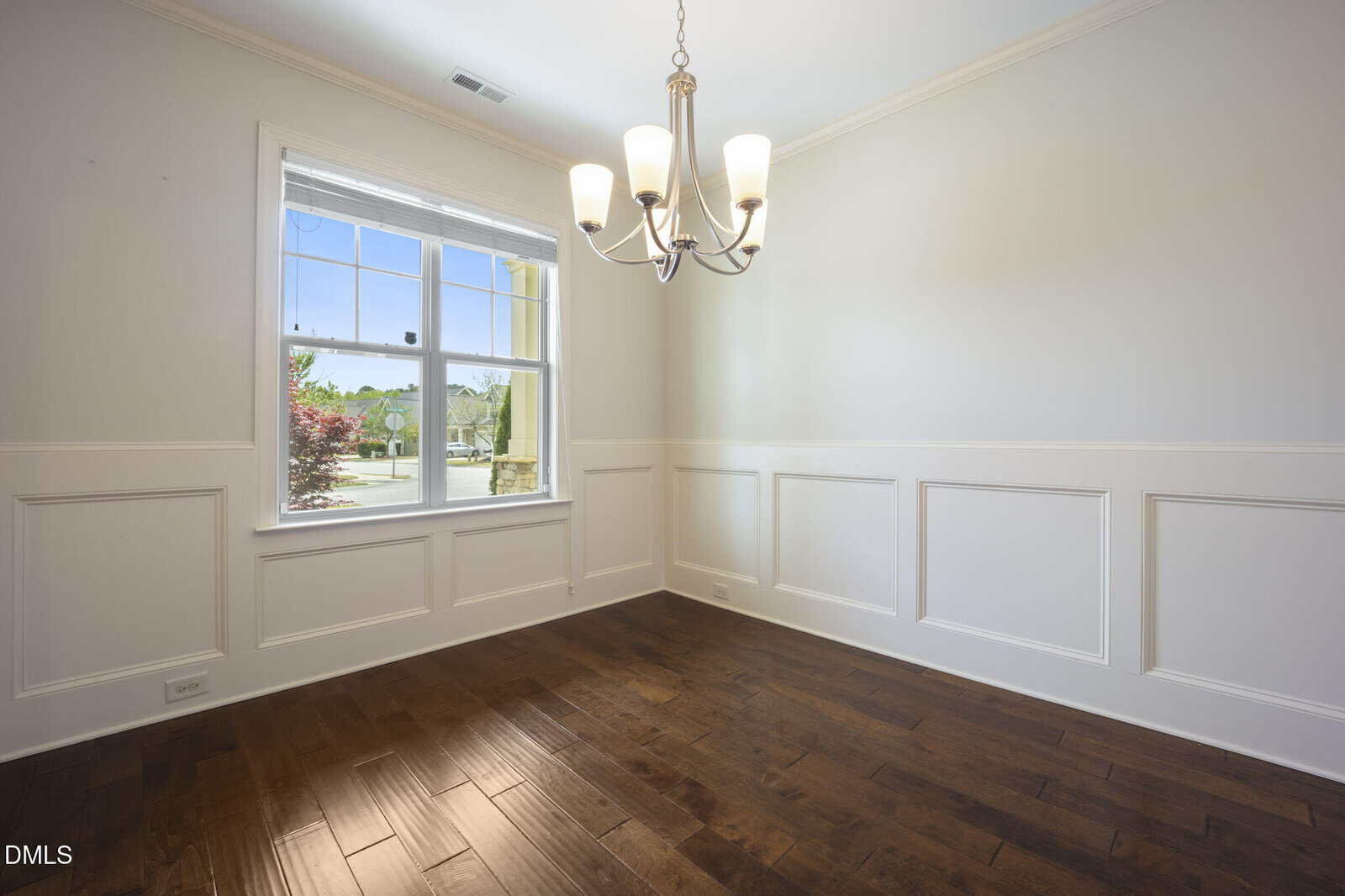 Empty room with white wainscoting, beige walls, a large window letting in natural light, dark hardwood floors, and a modern chandelier hanging from the ceiling.