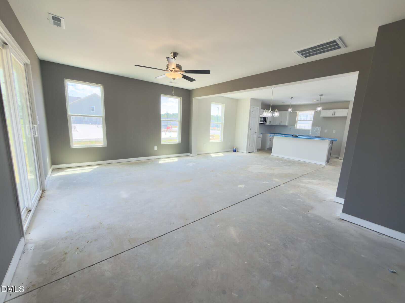 Spacious, unfurnished open living area with gray walls, large windows, ceiling fan, and visible kitchen with blue-taped island countertop. Light streams in from sliding glass doors and windows, and flooring is unfinished concrete.