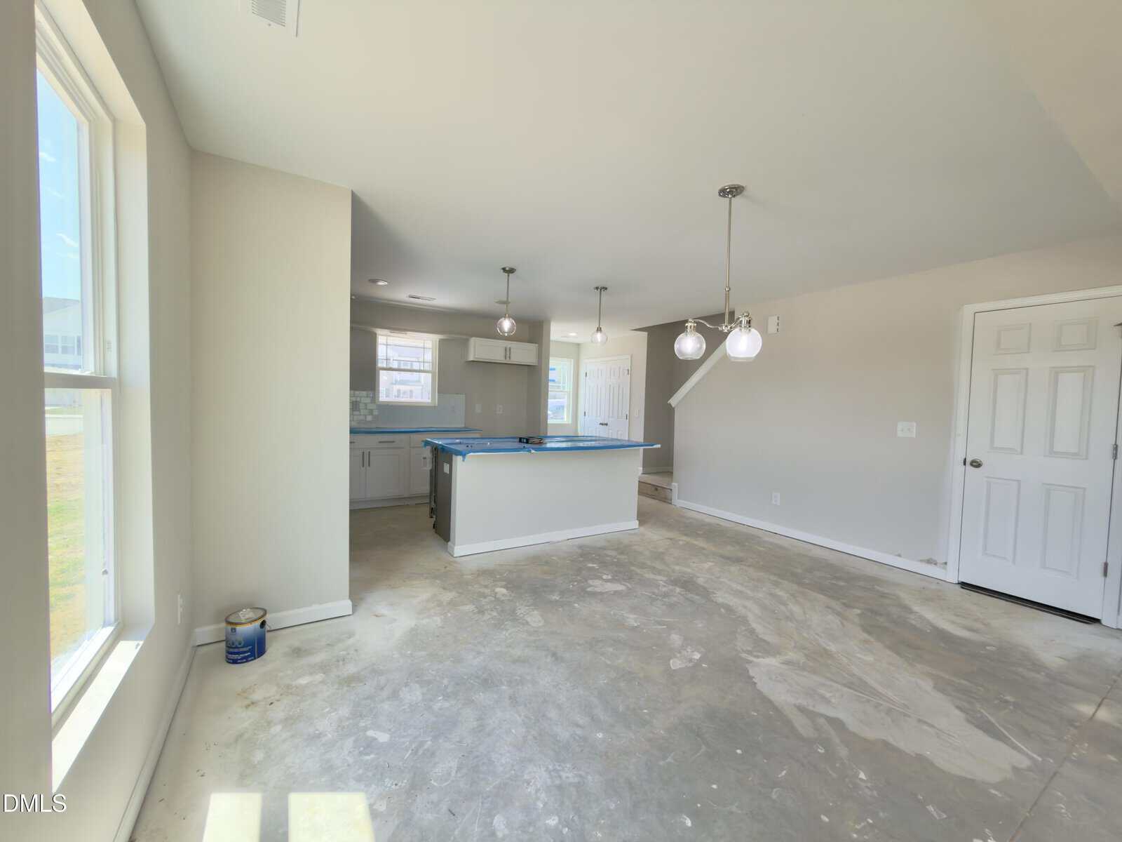 Unfinished kitchen and dining area with bare concrete floors, a central island covered by blue plastic, pendant lights above, white cabinets, and sunlight streaming through large windows on the left.