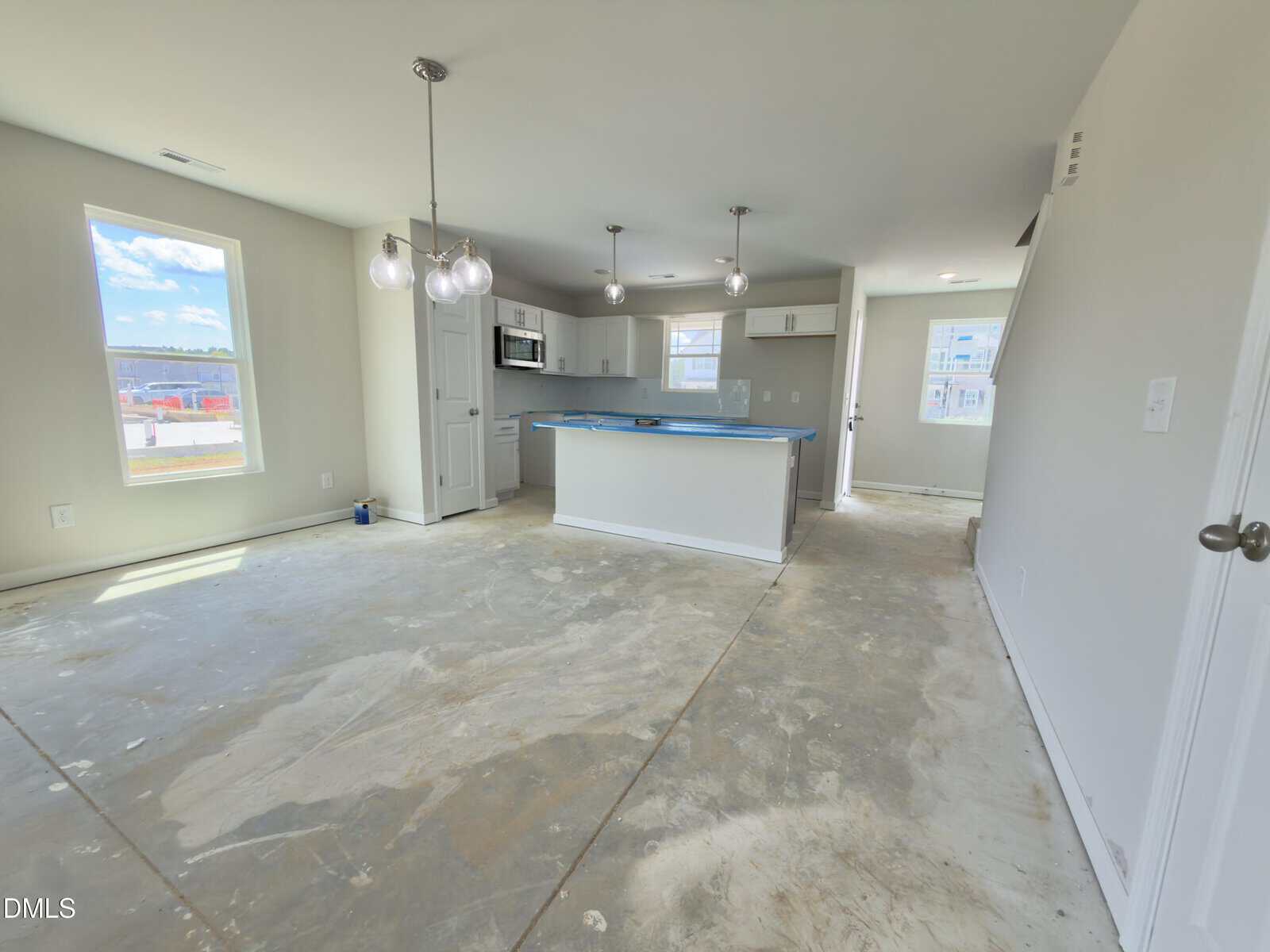 Unfinished open-concept kitchen and living area with exposed subfloor, white walls, and hanging light fixtures; sunlight streams through multiple windows.