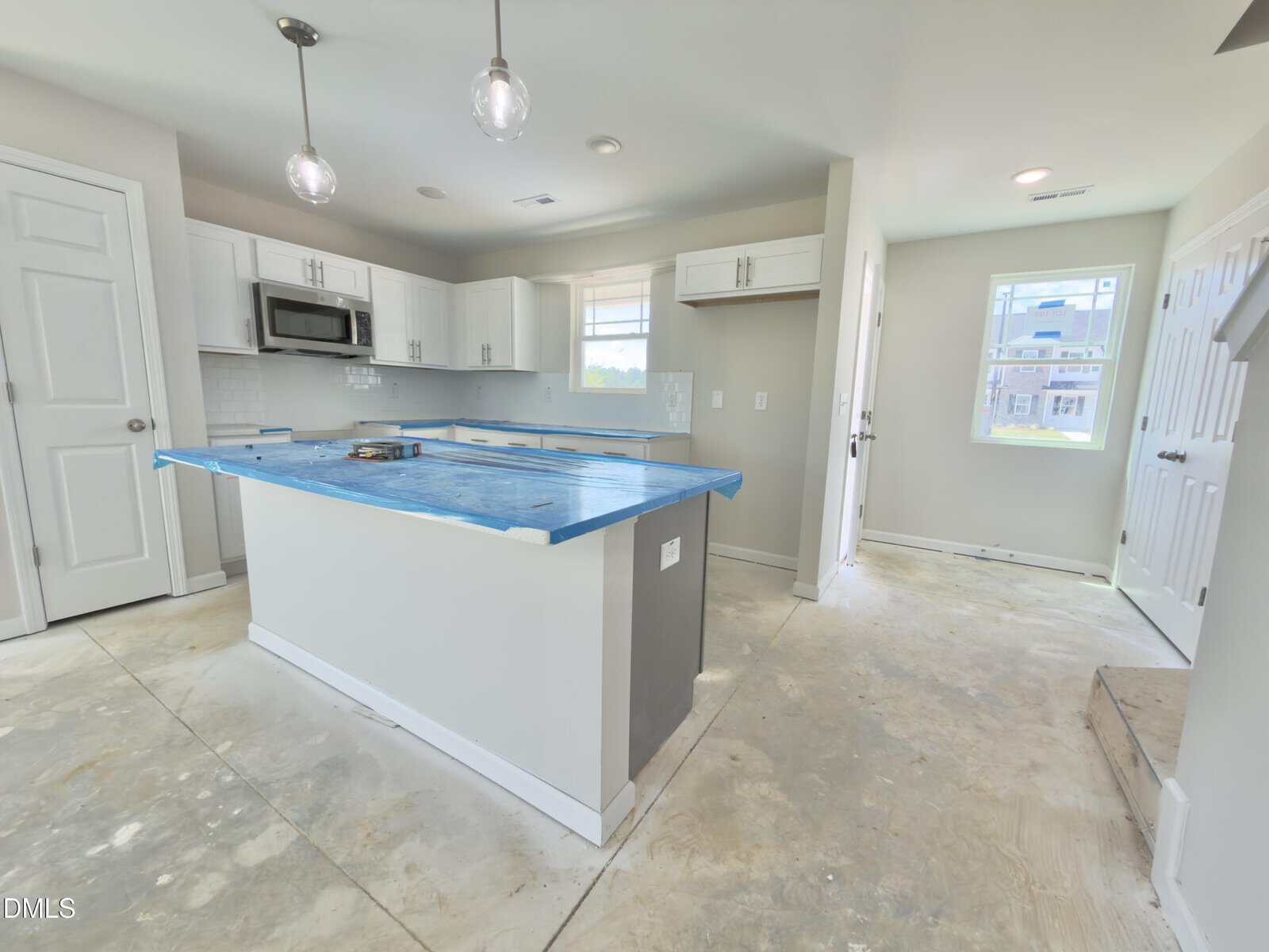 A newly constructed kitchen with white cabinets, a blue-covered island, pendant lights, and unfinished floors. There are two windows, a microwave above the stove, and multiple doors leading to other rooms.