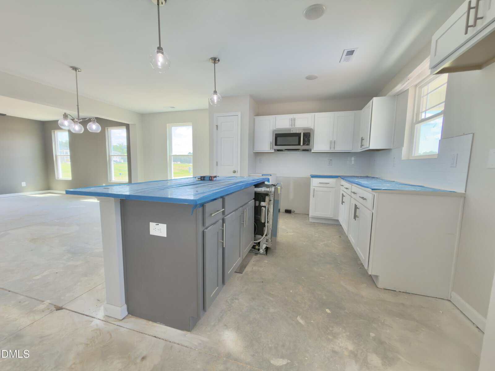 A modern kitchen with white cabinets, gray island, pendant lights, unfinished floors, and blue protective film on counters and appliances. Open layout connects to dining and living areas.