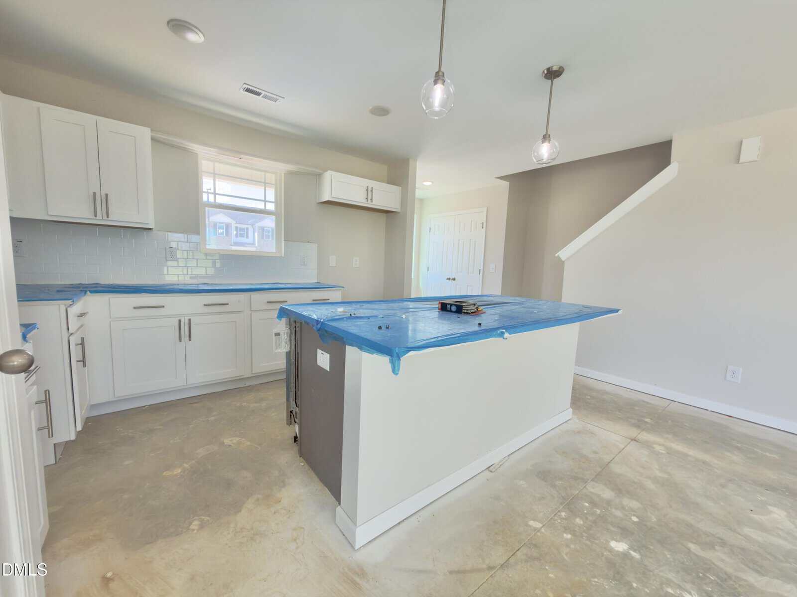 A kitchen under construction with unfinished floors, white cabinets, blue plastic covering the island, and two pendant lights hanging above. A staircase and closed door are visible in the background.