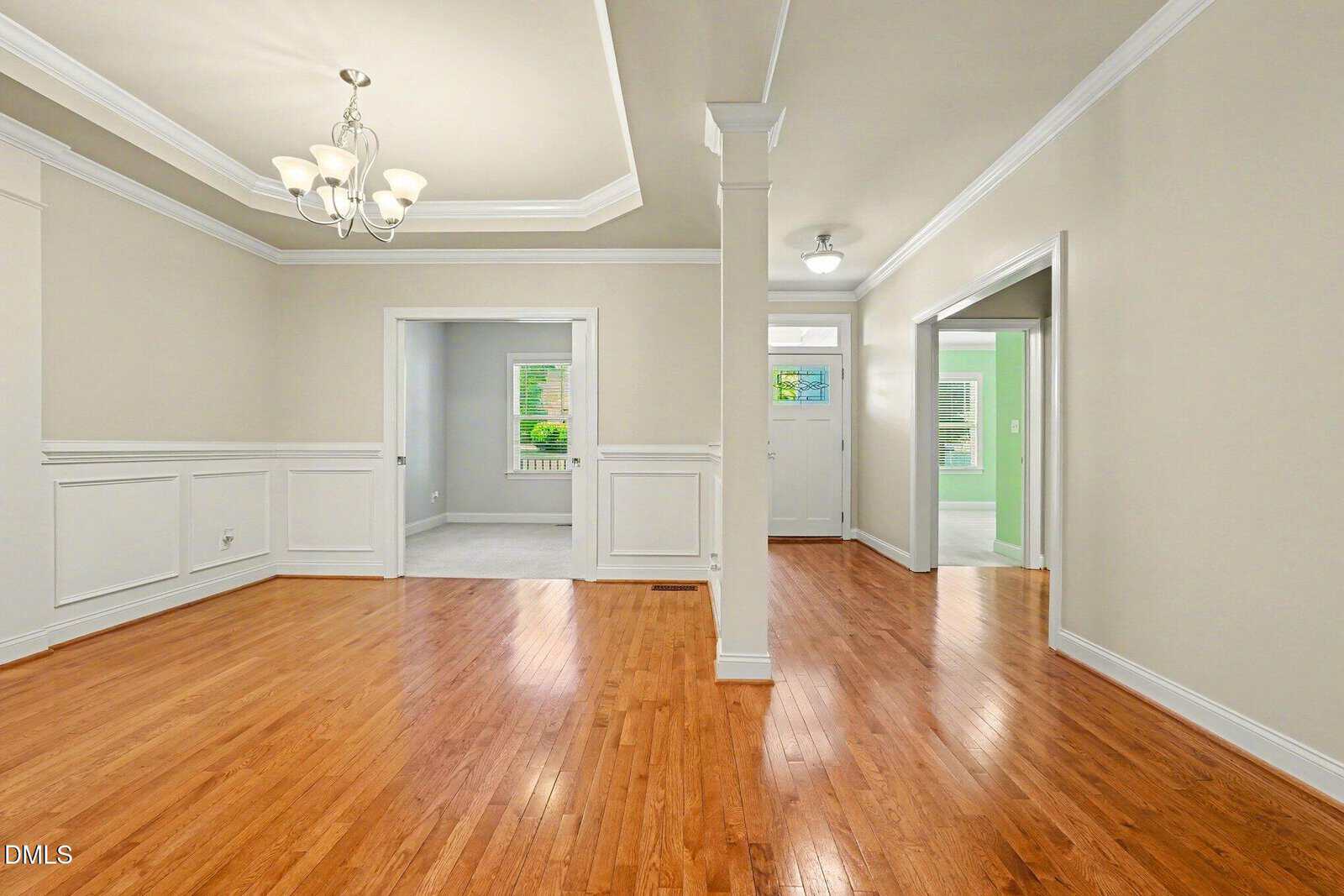 Bright, empty room with hardwood floors, beige walls, white trim, and a chandelier. Two open doorways lead to adjacent rooms; one room has green walls and the other features windows with outdoor views.