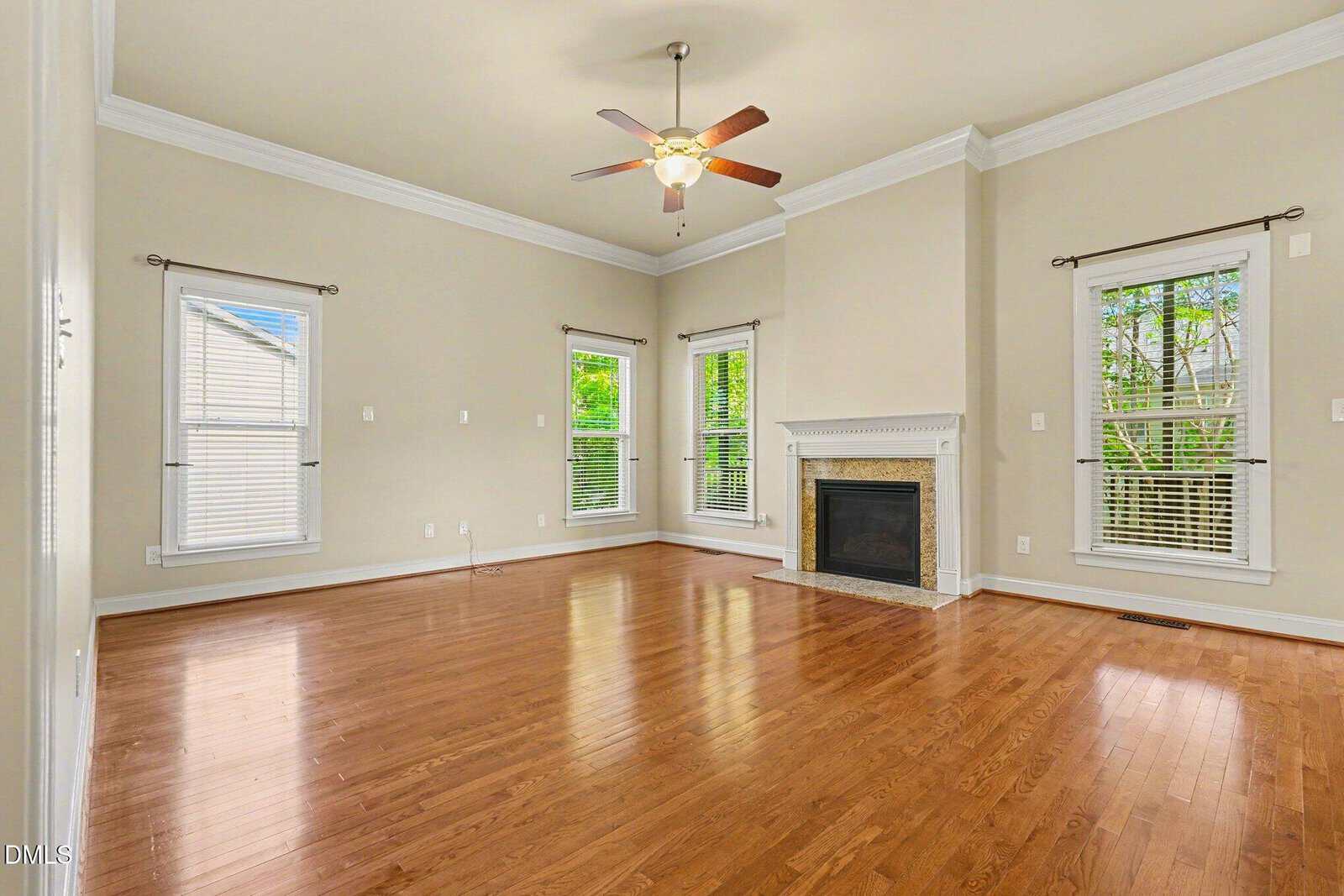 Bright living room with light beige walls, large windows, hardwood floors, white trim, a tray ceiling with ceiling fan, and a white fireplace with decorative mantel. No furniture is present.