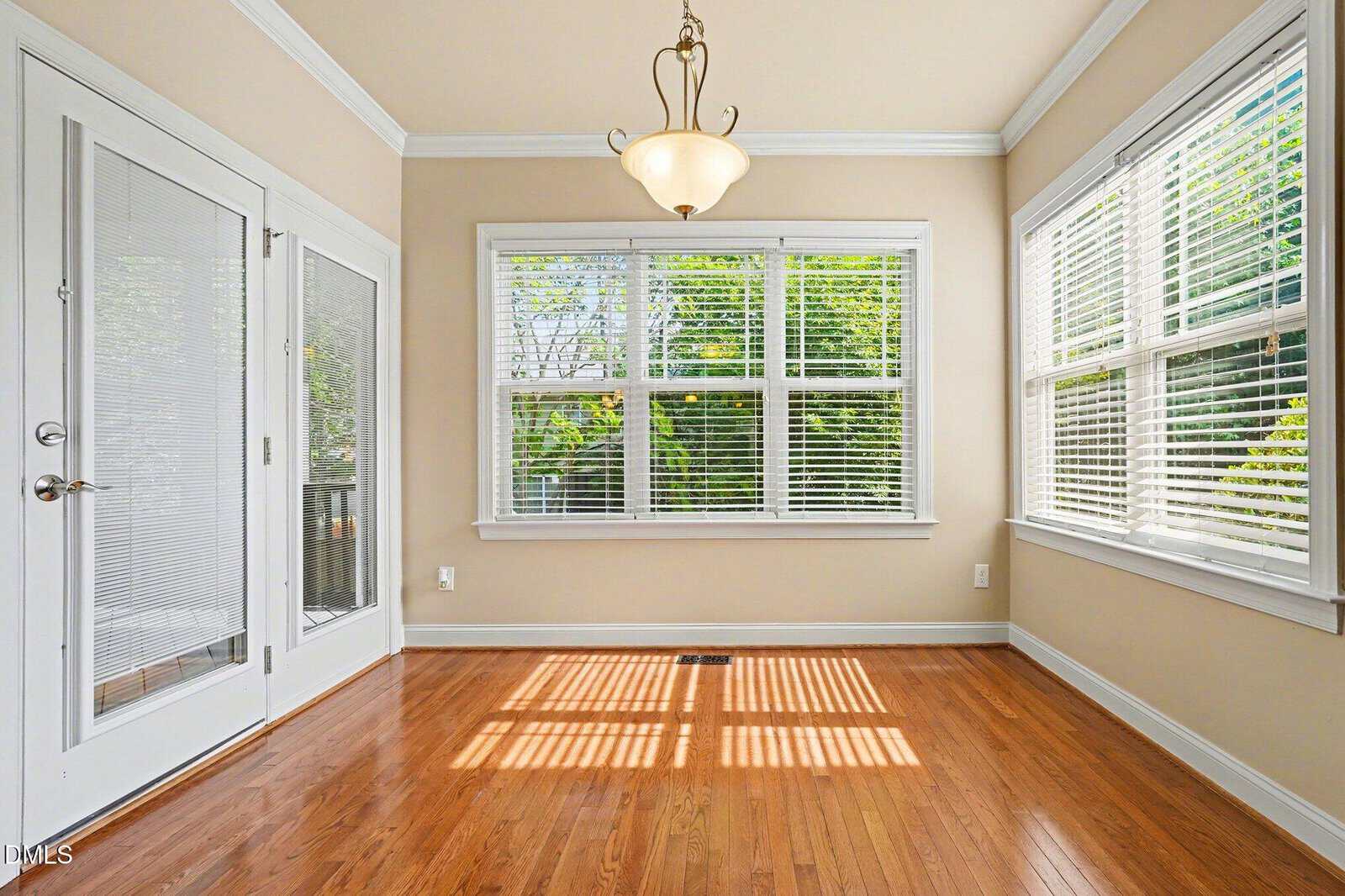 Bright sunroom with large windows, white blinds, beige walls, hardwood floors, and a hanging light fixture. Glass doors on the left open outside. Sunlight creates a square pattern on the floor.