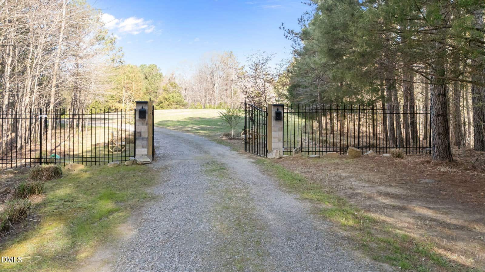 A gravel driveway leads through an open black iron gate with stone pillars, surrounded by trees and grassy areas on a clear, sunny day.