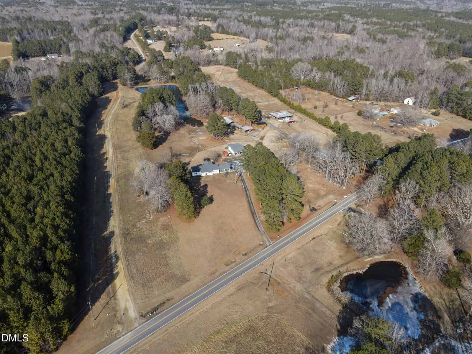 Aerial view of a rural landscape with a house surrounded by trees, open fields, a pond, a road running through the area, and additional houses and wooded areas in the background.