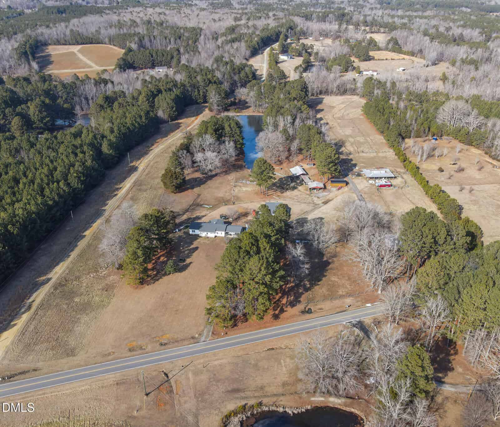 Aerial view of a rural property featuring multiple buildings, a pond, open fields, large trees, and a road in the foreground, with wooded areas and additional structures in the background.