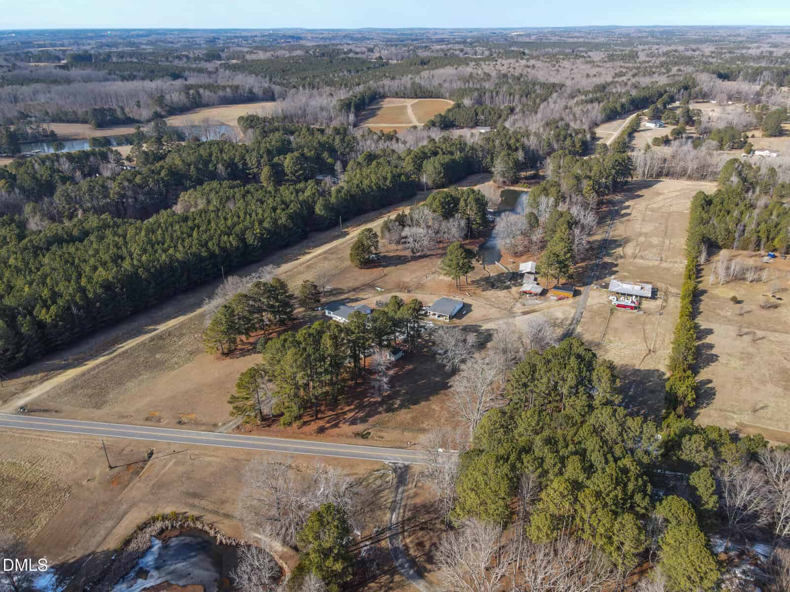 Aerial view of a rural area with a road running through a landscape of trees, open fields, and several houses scattered among wooded sections and clearings. The scene appears calm and sparsely populated.