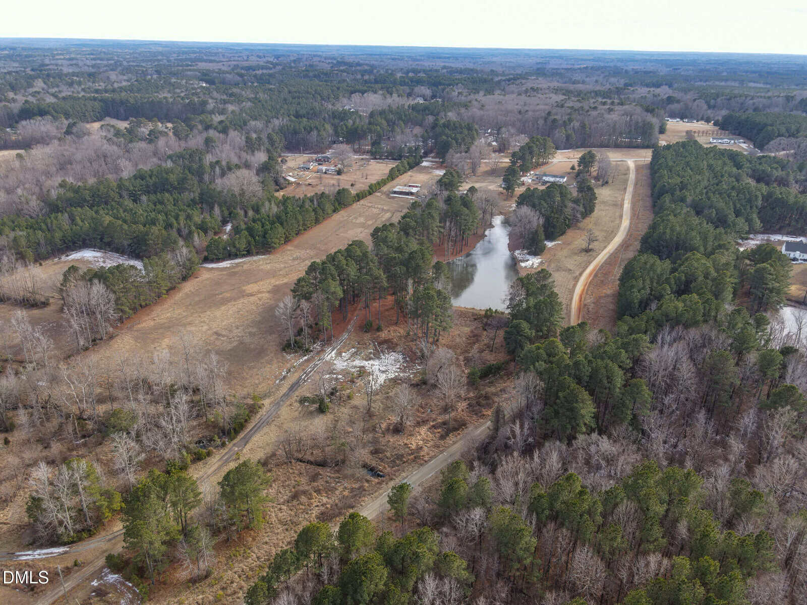 Aerial view of a rural landscape with a pond, open fields, dense clusters of trees, and a winding road passing through the area. Several structures are visible in the distance among the trees.