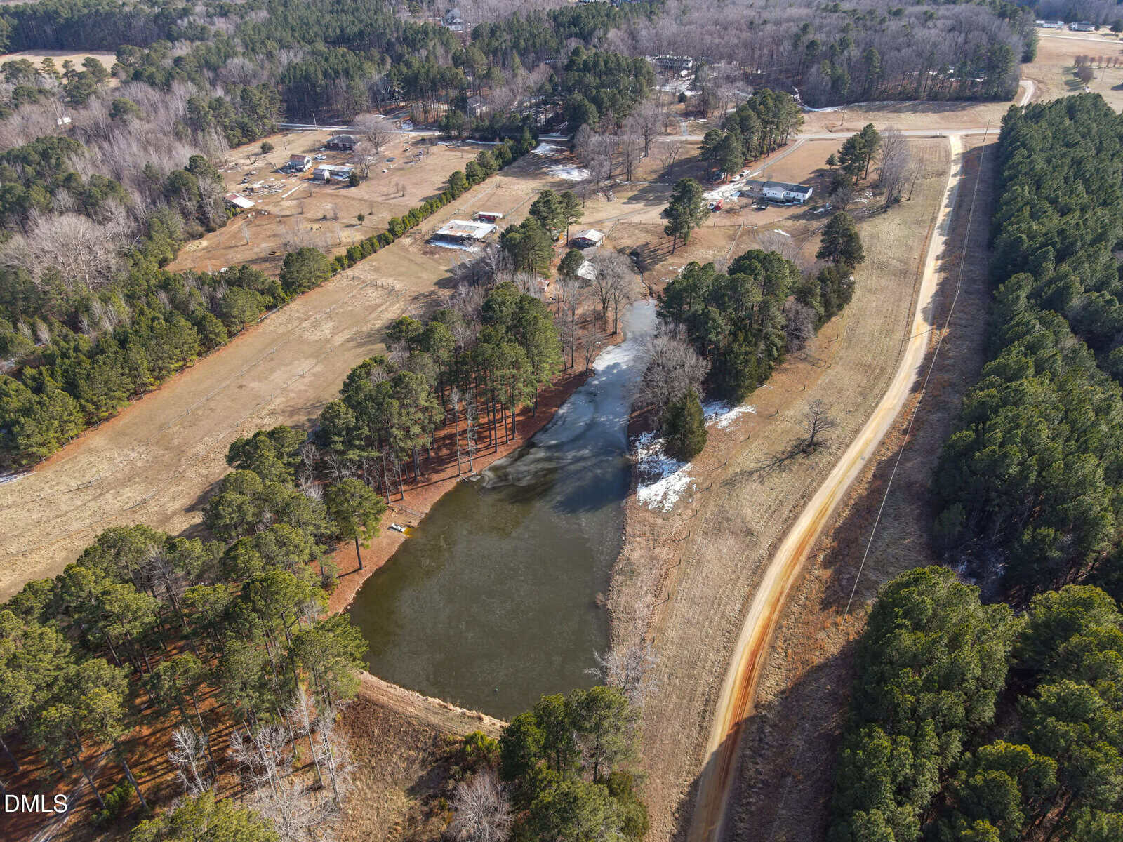 Aerial view of a rural landscape featuring a pond surrounded by trees, dirt roads, scattered houses, and open fields. Some areas have patches of snow or ice near the pond and along the wooded areas.