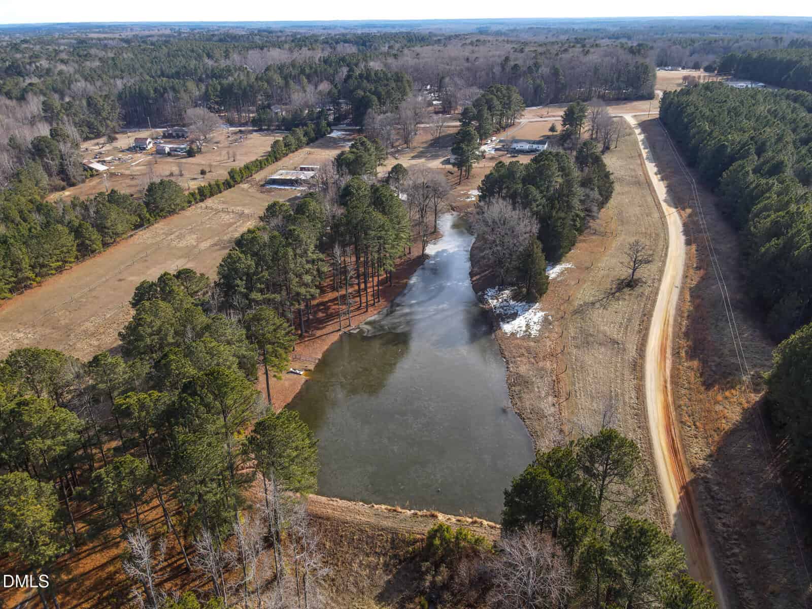 Aerial view of a rural landscape with a pond surrounded by trees, a dirt road running alongside, and scattered homes and farmland in the background under a clear sky.