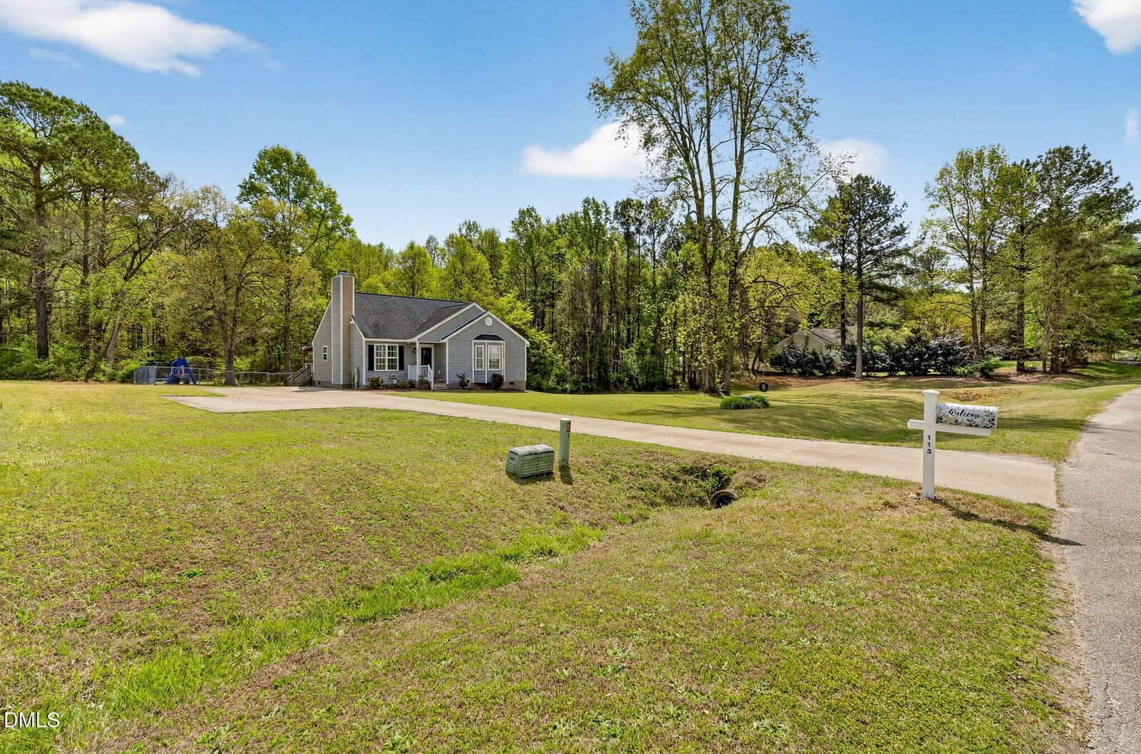 A single-story gray house with white trim and a chimney sits on a large grassy lot with a paved driveway. A white mailbox and a white post are near the roadside, and trees surround the property under a blue sky.