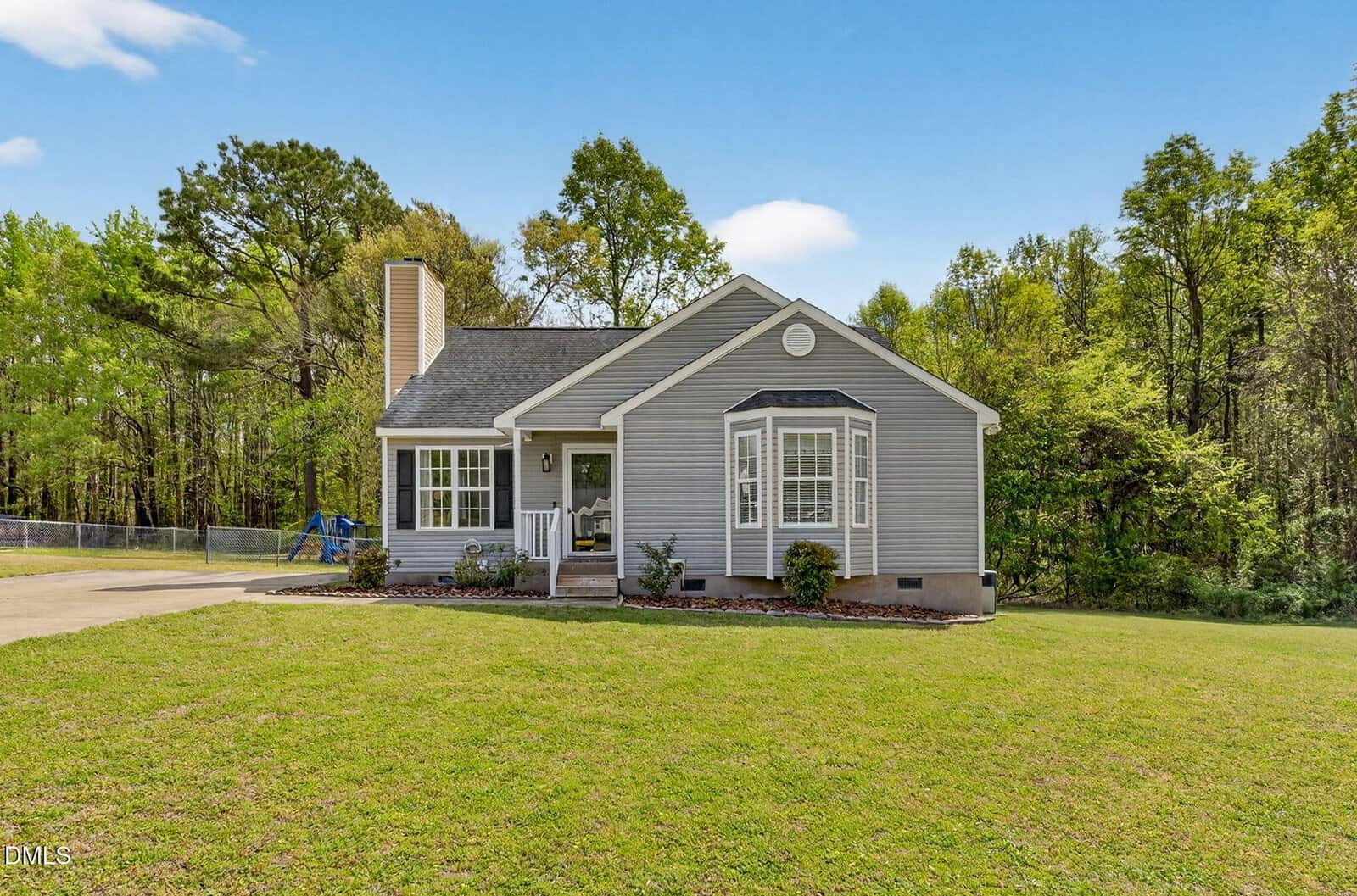 A single-story gray house with white trim sits on a large, grassy lawn. There are a few shrubs by the covered front porch, and the house is surrounded by trees under a clear blue sky.