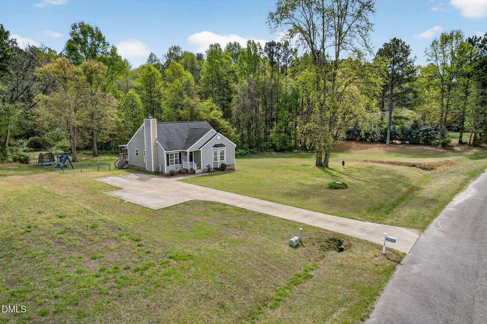 A small, single-story gray house with white trim sits on a large grassy lot with a long driveway, surrounded by trees; a swing set is visible in the backyard near a wooded area.