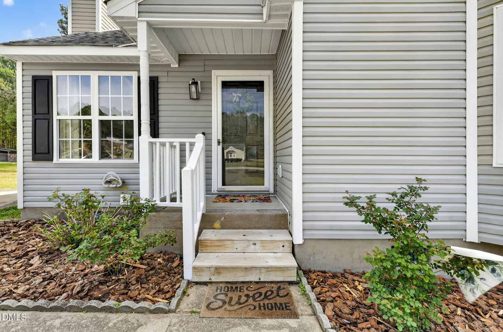 A small set of wooden steps leads to a glass front door of a gray house with white trim. A doormat with "Home Sweet Home" is at the entrance, and there are green bushes and mulch along the walkway.