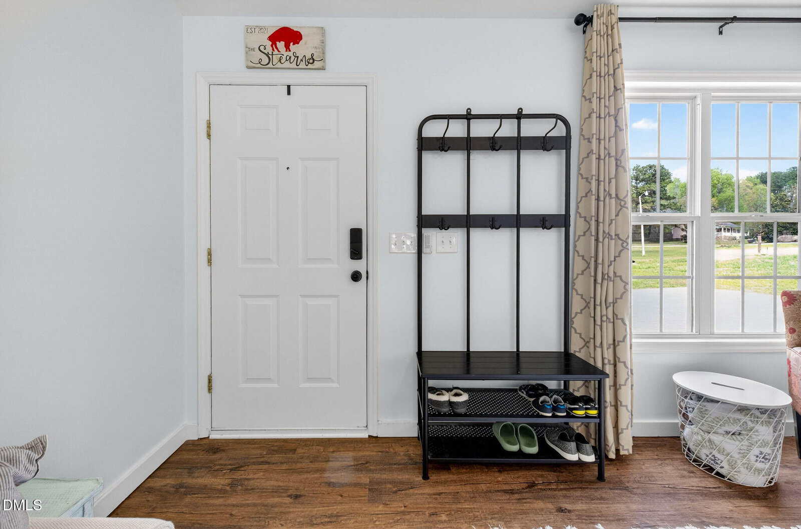 A white front door next to a black metal shoe rack and coat hanger with several pairs of shoes. A window with beige curtains lets in daylight. A decorative sign hangs above the door on a white wall.