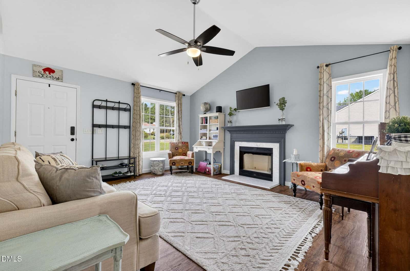Bright living room with vaulted ceiling, beige sofa, fireplace, TV, two windows, armchairs, rug, piano, and ceiling fan. Decor includes plants, shelves, and wall art. Natural light fills the space.