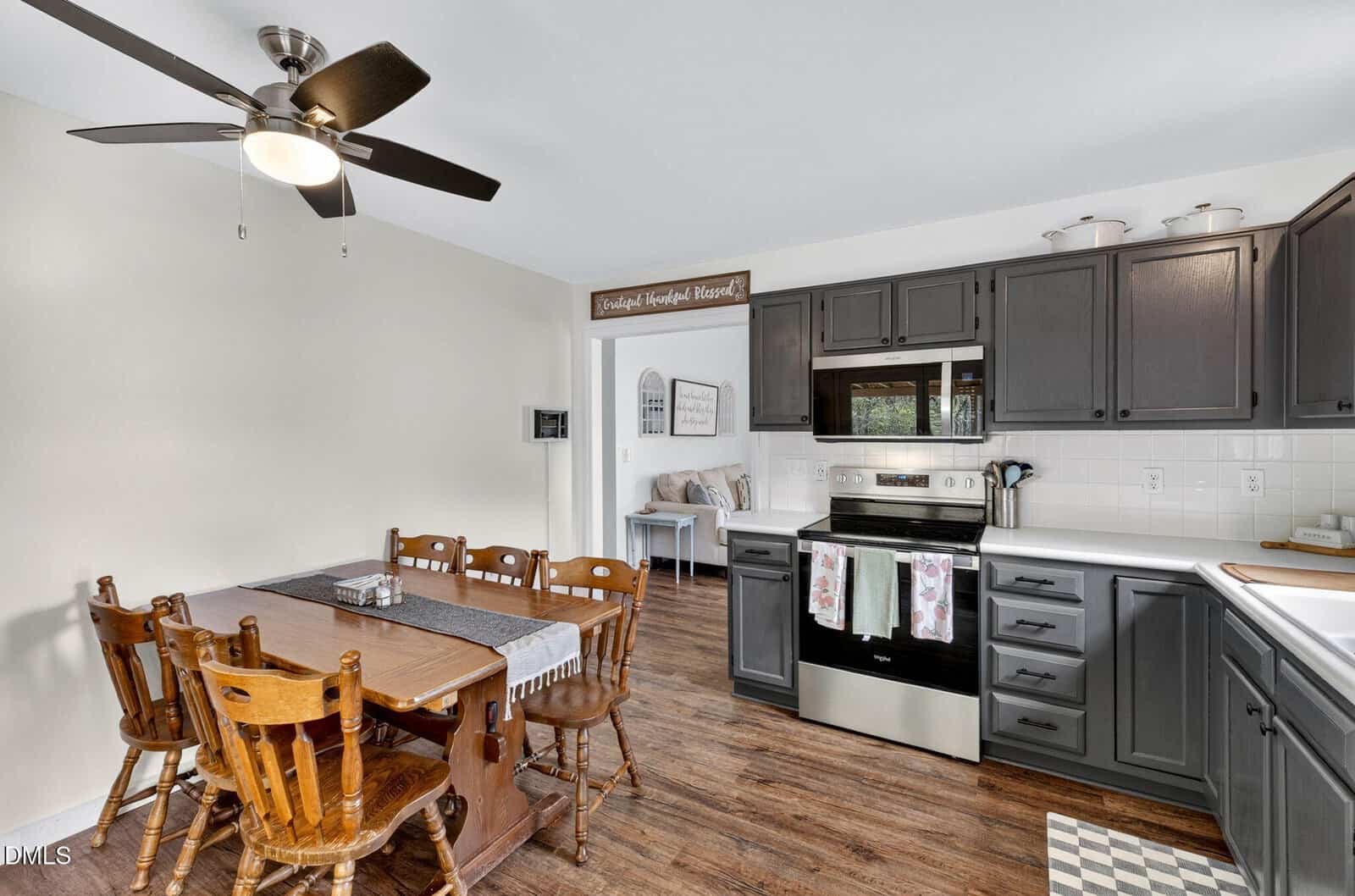 Kitchen with dark cabinets, white countertops, electric stove, and dining area with a wooden table and four chairs. Ceiling fan above, wood flooring, and farmhouse-style decor visible throughout the space.