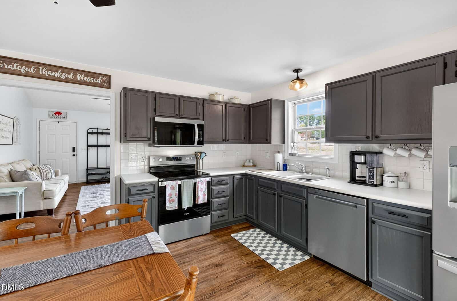 Modern kitchen with gray cabinets, white countertops, stainless appliances, and a window over the sink. Dining table in the foreground, view into a living room with a sofa in the background. Bright, clean, and organized space.
