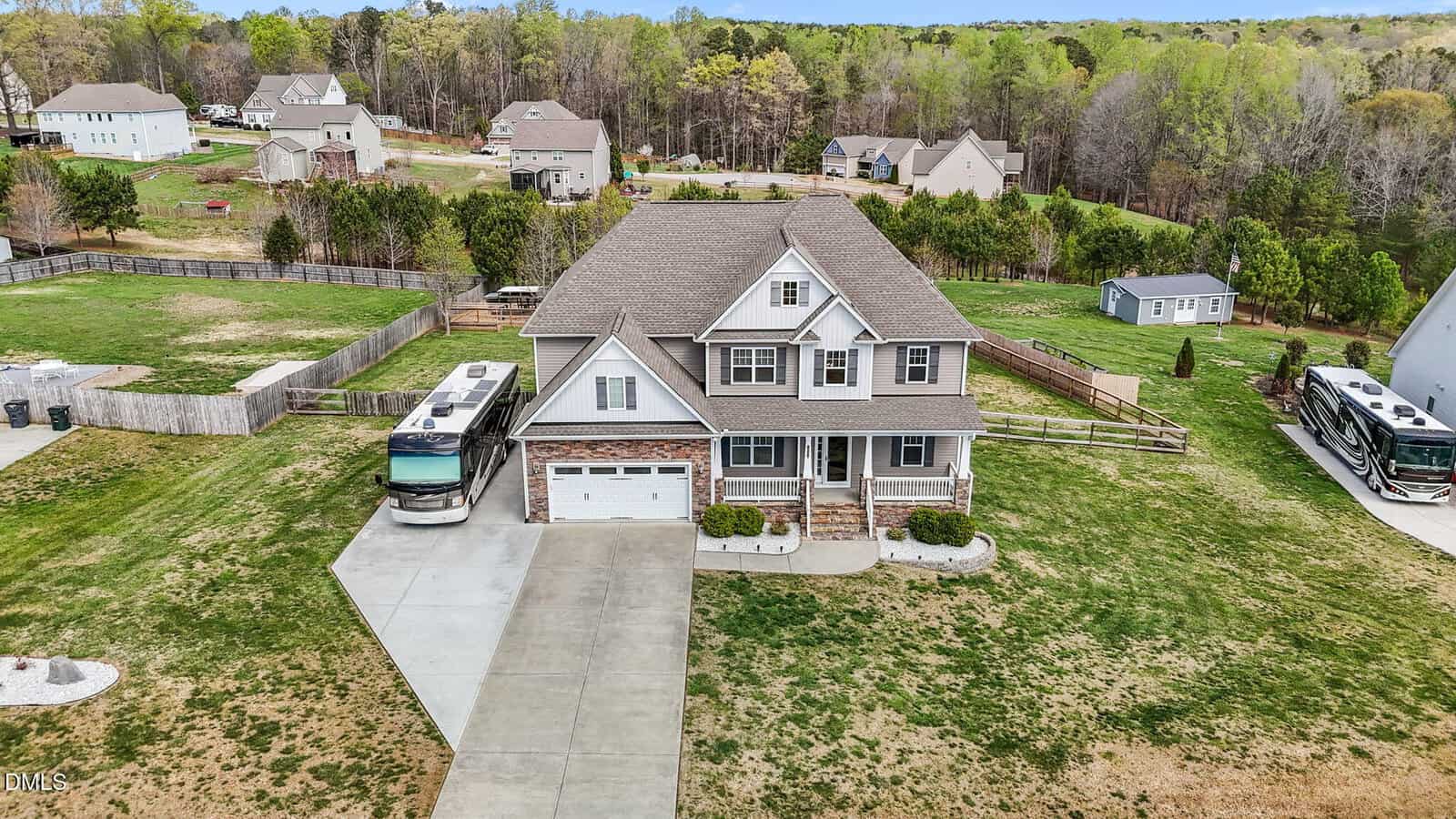 Aerial view of a two-story suburban house with a large driveway, garage, front porch, and neatly trimmed lawn. An RV is parked beside the house, and neighboring houses with spacious yards are visible in the background.