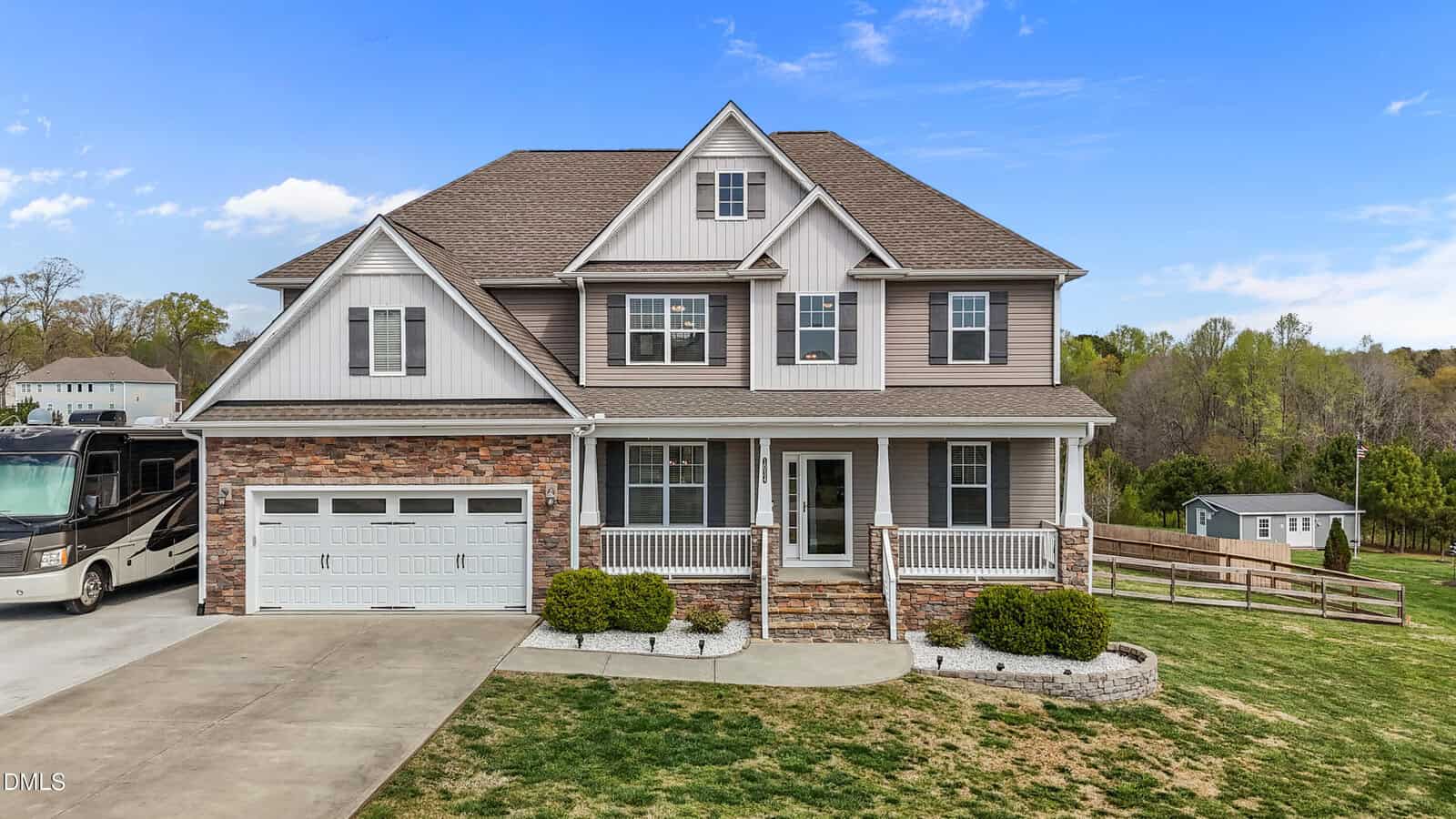 A two-story suburban house with a front porch, attached double garage, and stone accents. The home sits on a spacious lawn with a driveway, a small shed, and an RV parked nearby under a blue sky with clouds.