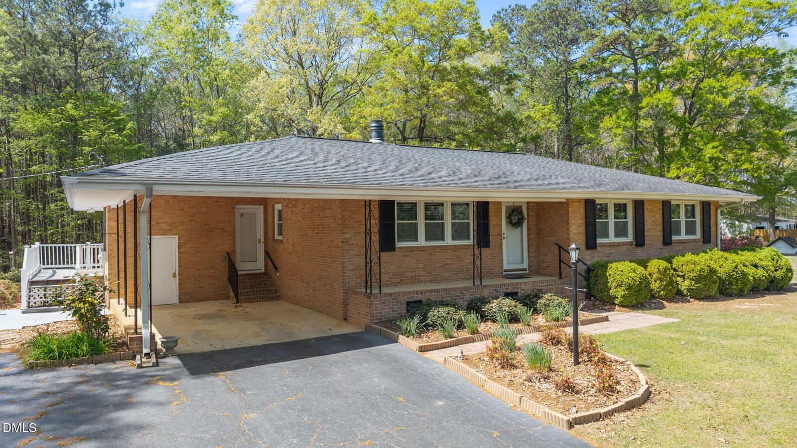 Single-story brick house with a dark shingle roof, covered carport on the left, black iron posts, and a landscaped front yard with shrubs and a curved flower bed. Surrounded by tall trees and greenery.