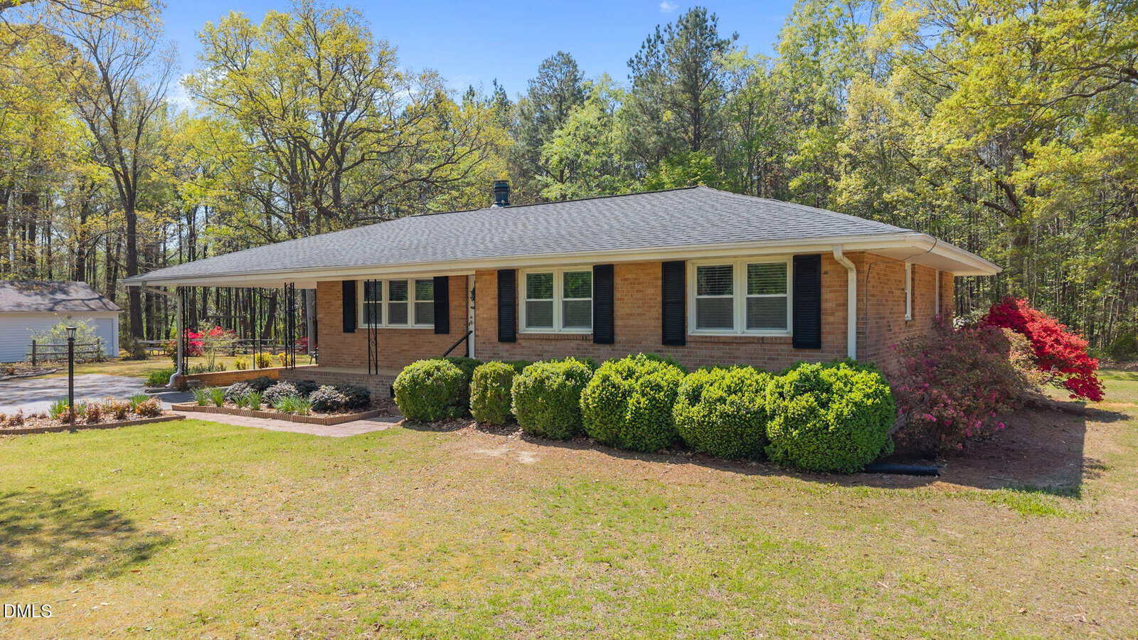 A single-story brick house with a gray shingle roof, black shutters, and white trim, surrounded by trimmed bushes and trees. The yard is grassy with some flowering plants and a wooded area in the background.