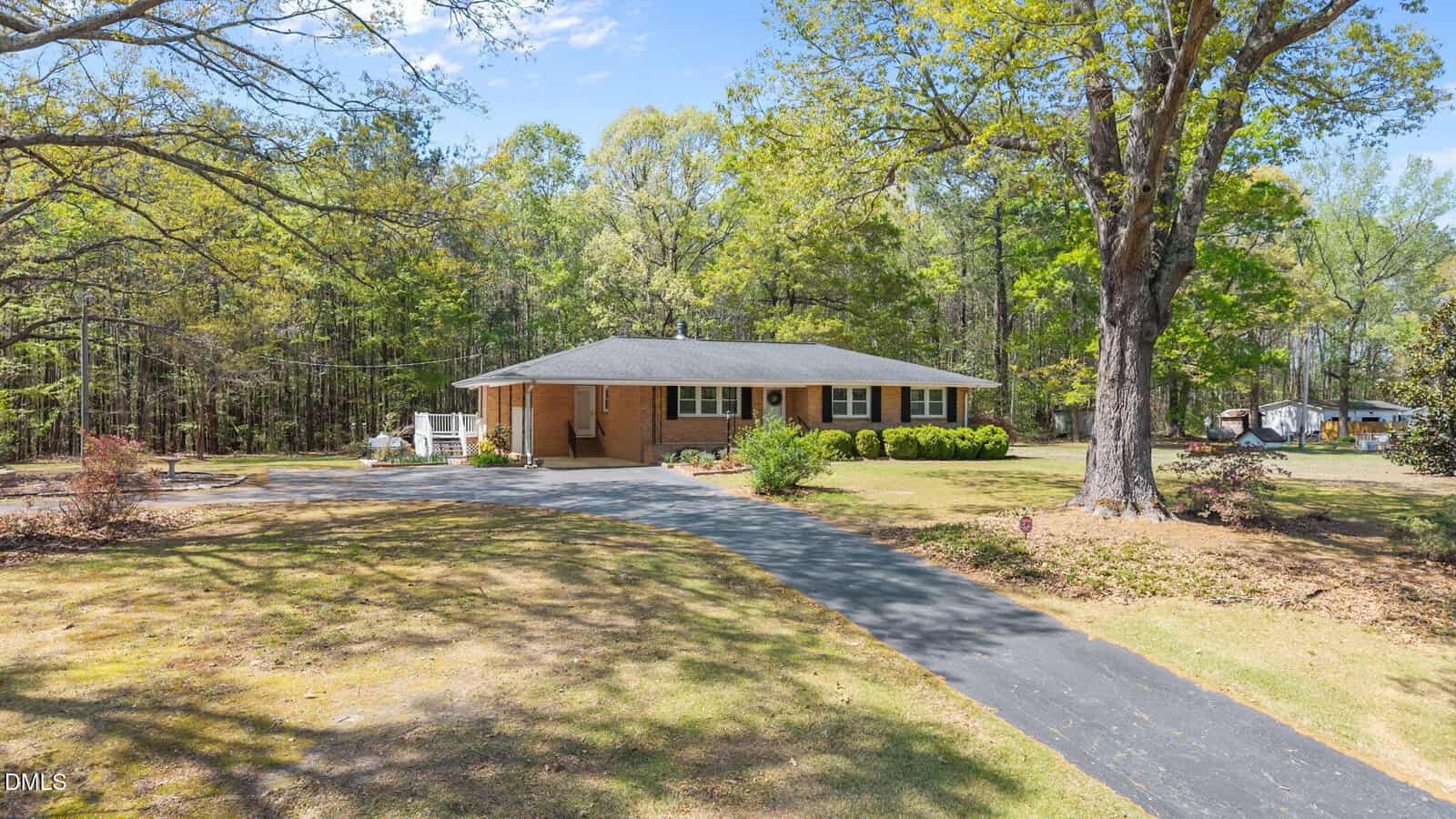Single-story brick house with a front porch, surrounded by trees and bushes. A curved driveway leads to the house, and the yard has patches of grass and dry leaves. The background shows dense woods and a partly cloudy sky.