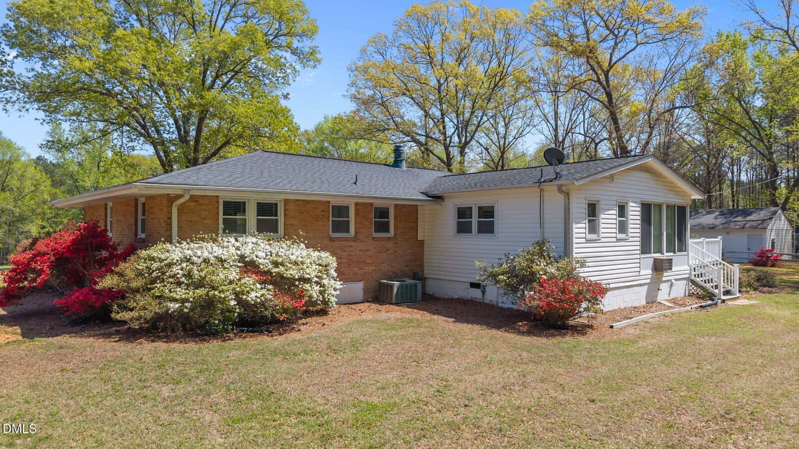 A single-story house with a brick and white exterior sits on a grassy yard, surrounded by blooming bushes and tall trees under a clear blue sky. An outdoor AC unit and satellite dish are visible on the side of the house.