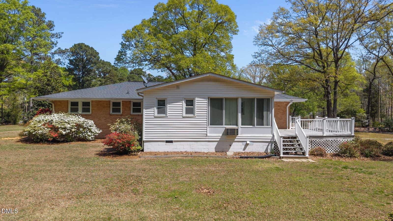 Single-story brick house with a white sunroom and deck, surrounded by green grass, blooming bushes, and tall trees under a clear blue sky.