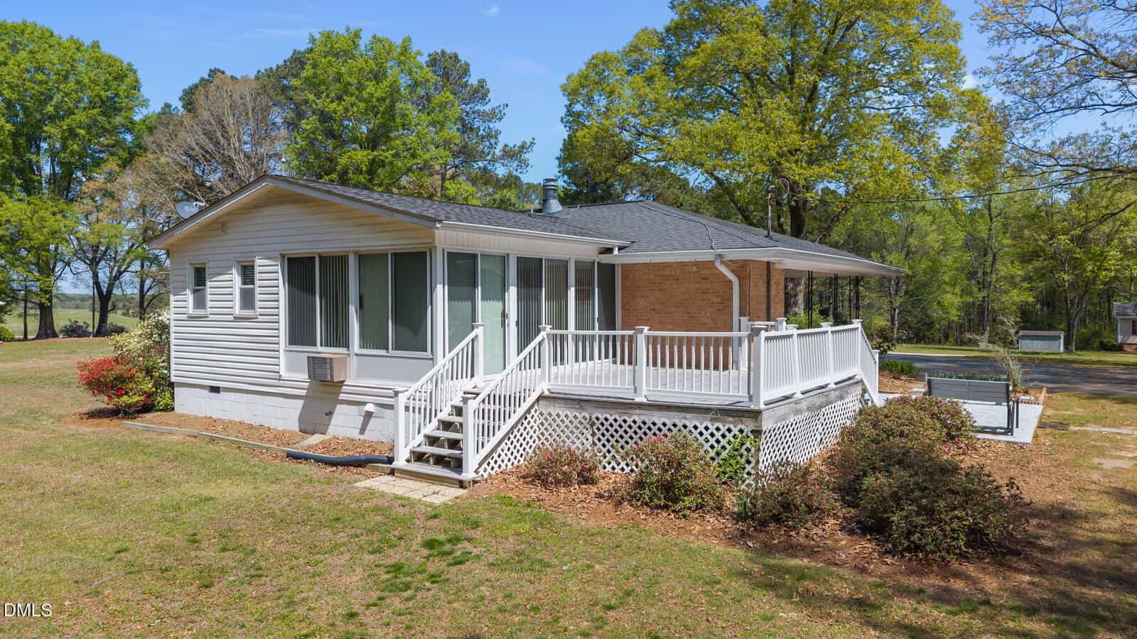 A small, single-story house with white siding, a brick accent wall, and a spacious wraparound porch with white railings, surrounded by trees and grass under a clear blue sky.