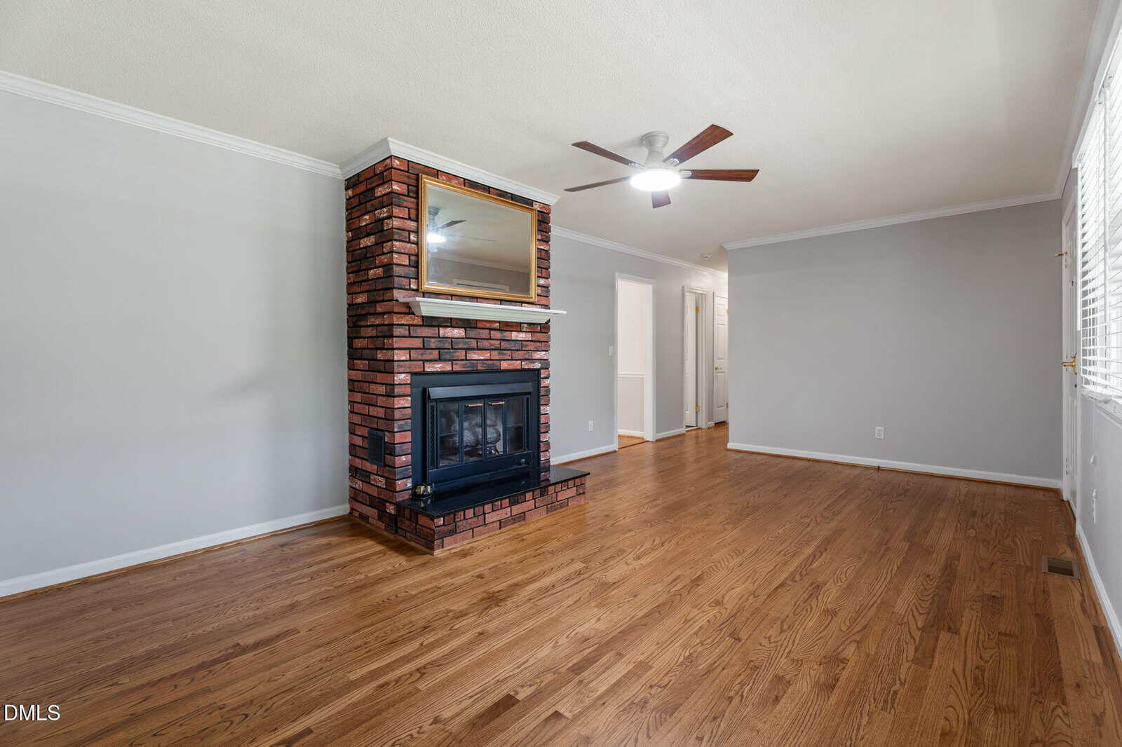 A spacious living room with hardwood floors, a brick fireplace, a ceiling fan with lights, light gray walls, and white trim. Large windows let in natural light.