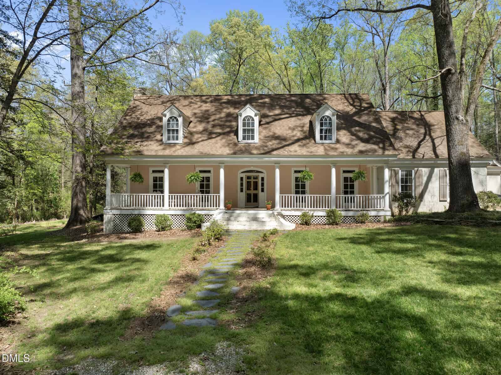 A two-story house with a tan exterior, white trim, and a wraparound front porch. The porch has hanging plants, and the house sits on a grassy lawn surrounded by tall trees. A stone path leads to the front steps.