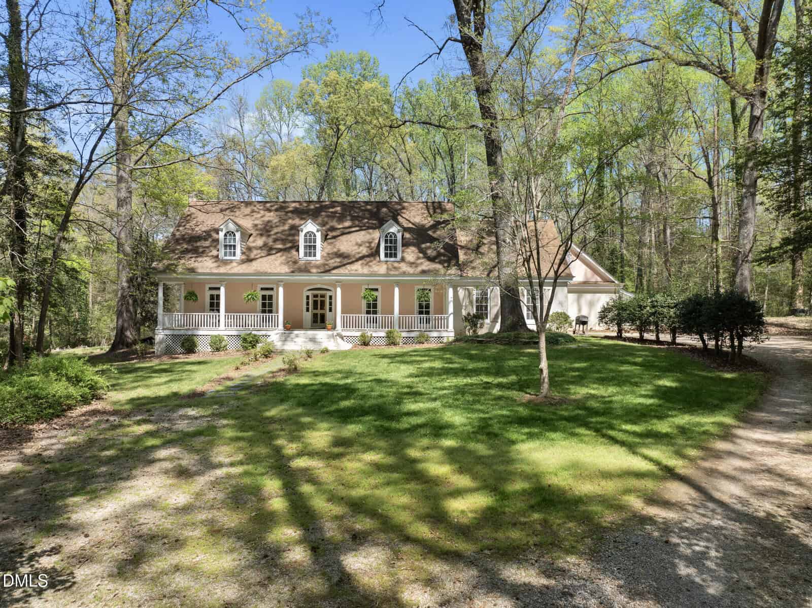 A large beige house with white trim and a front porch is surrounded by tall trees and green lawn. A gravel driveway curves to the right, and sunlight filters through the foliage.