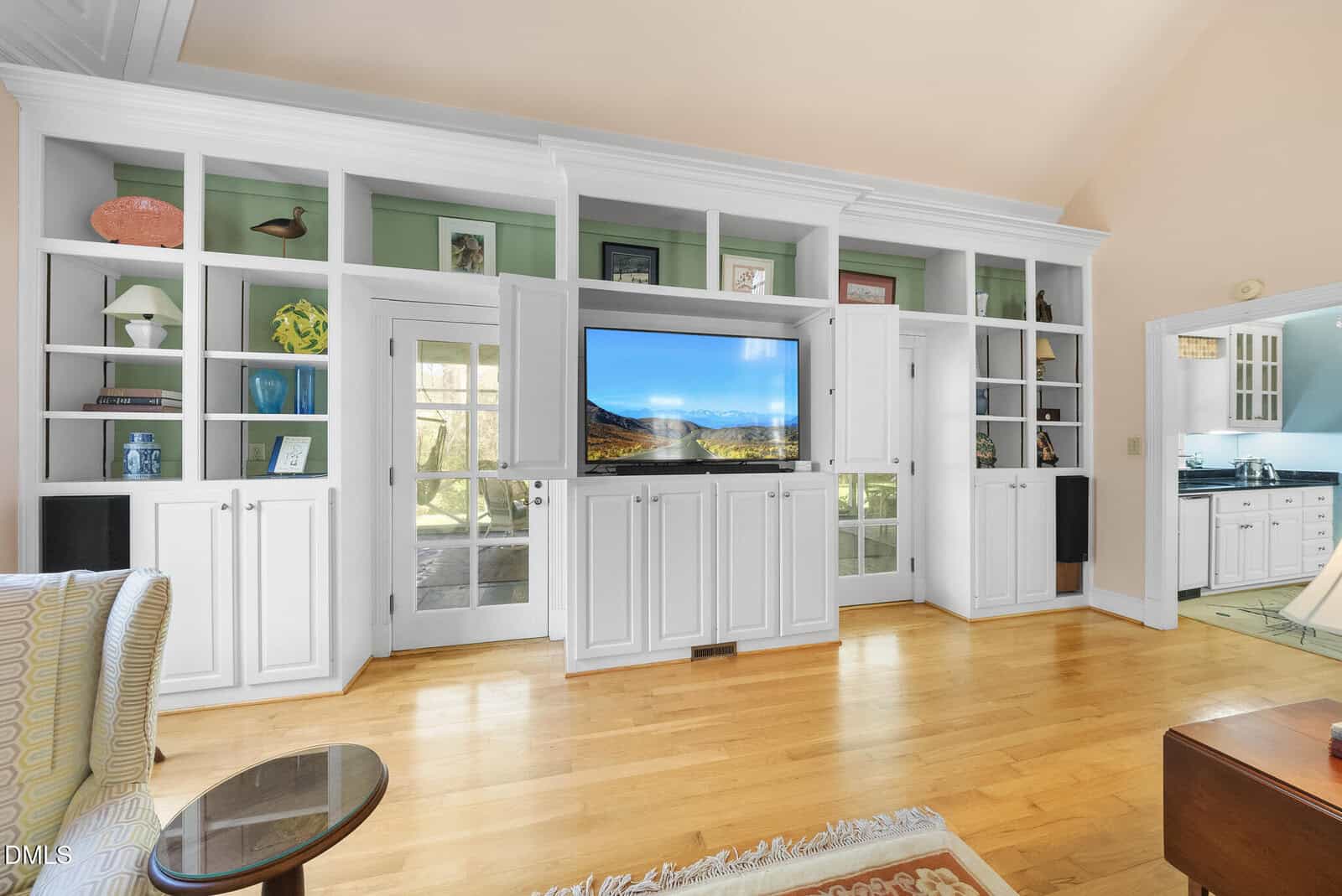 Built-in white shelves and cabinets line a living room wall, with a flat-screen TV at the center displaying a mountain scene. French doors and glass panels let in natural light. The room has wood floors and views into a kitchen.