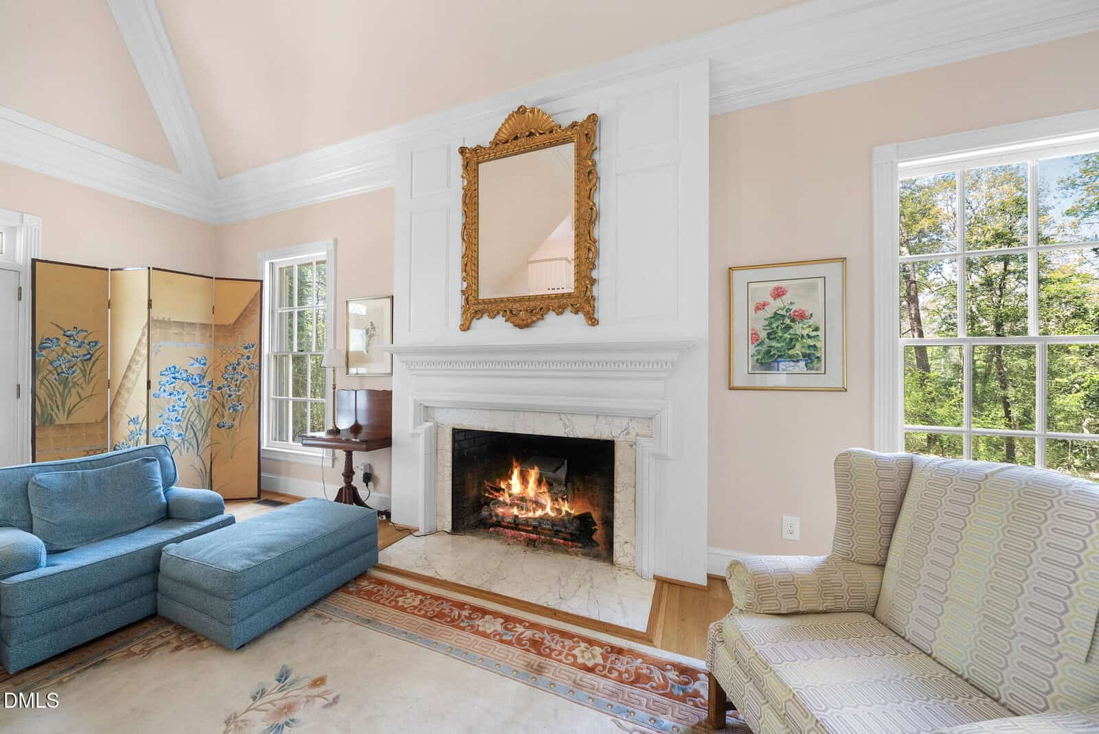 Elegant living room with a lit fireplace, large ornate mirror above the mantel, pastel walls, blue sectional, patterned armchair, floral art, large windows with greenery outside, and a decorative folding screen.
