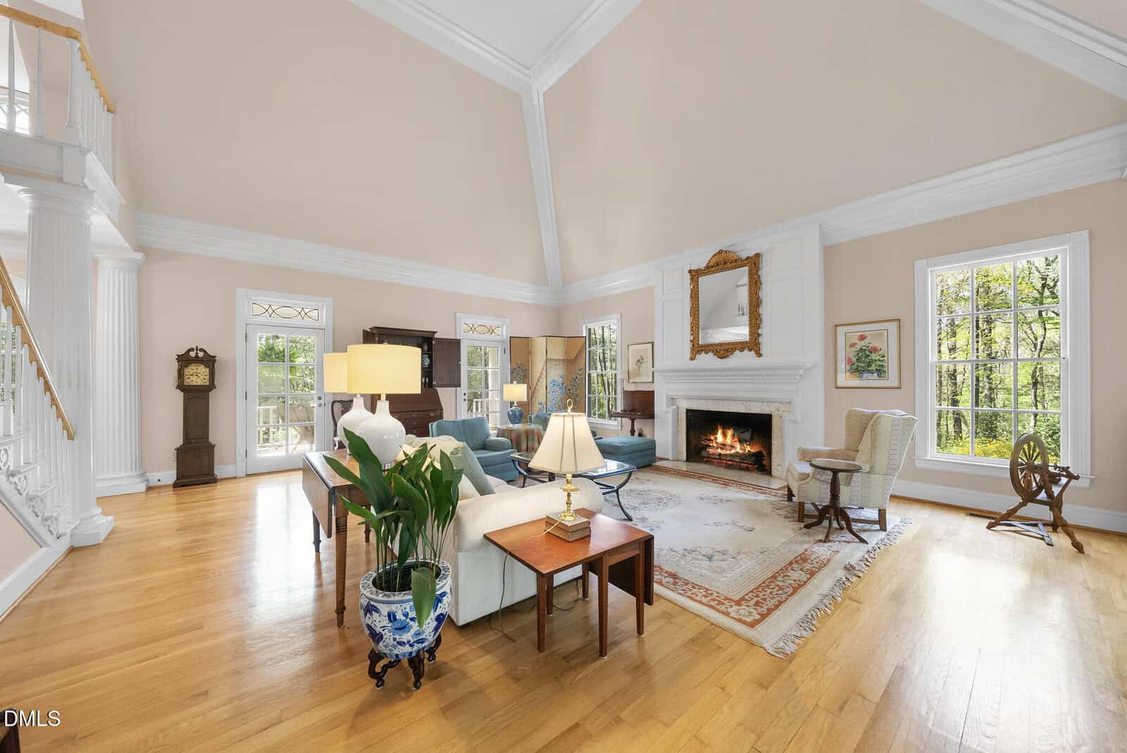 Spacious living room with high ceilings, hardwood floors, a fireplace, large windows, elegant furniture, a grandfather clock, and decorative plants, filled with natural light.
