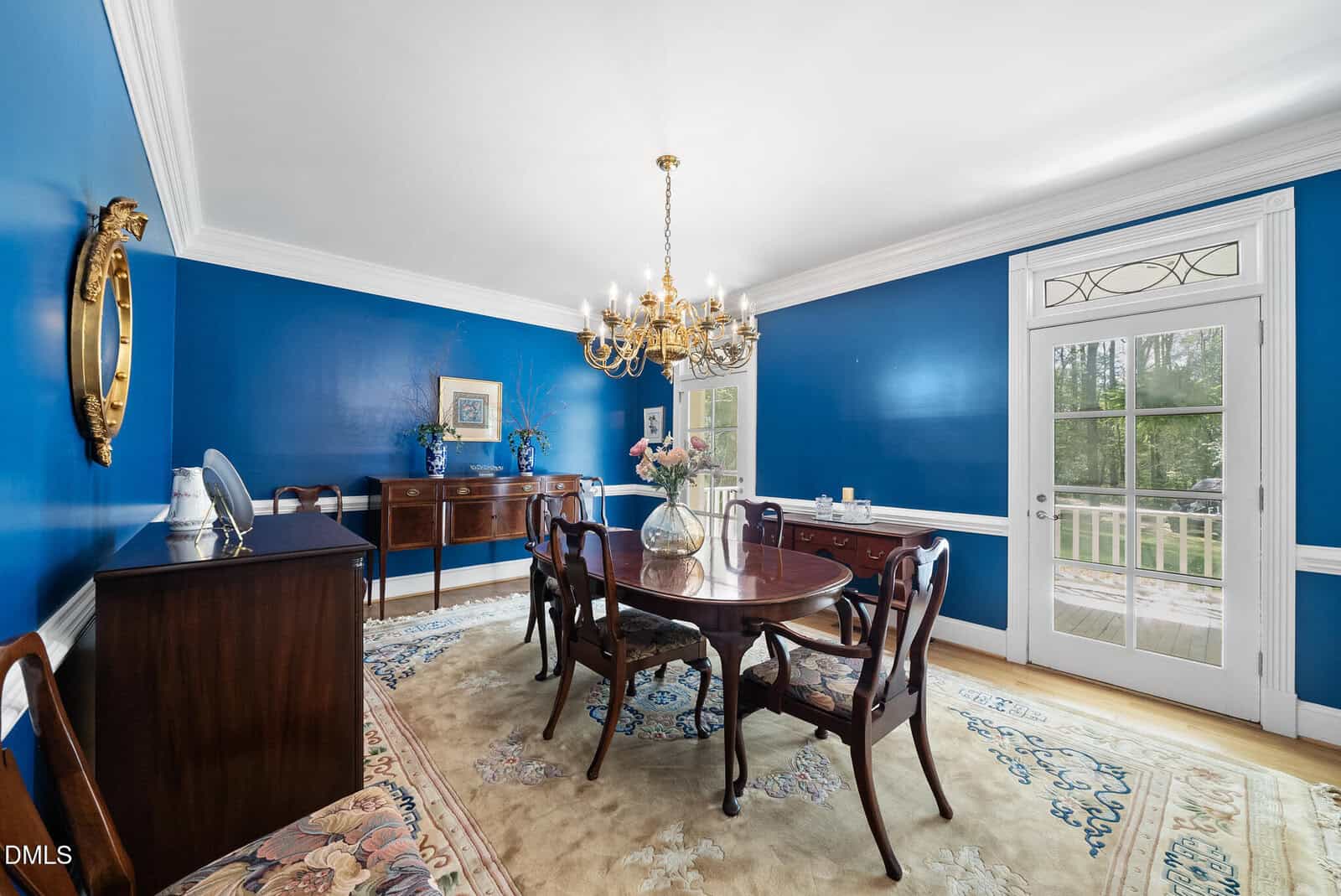 A traditional dining room with bright blue walls, a wooden dining table and chairs, a chandelier, sideboards, a large rug, and a door with glass panes leading outside. A mirror and framed art hang on the walls.