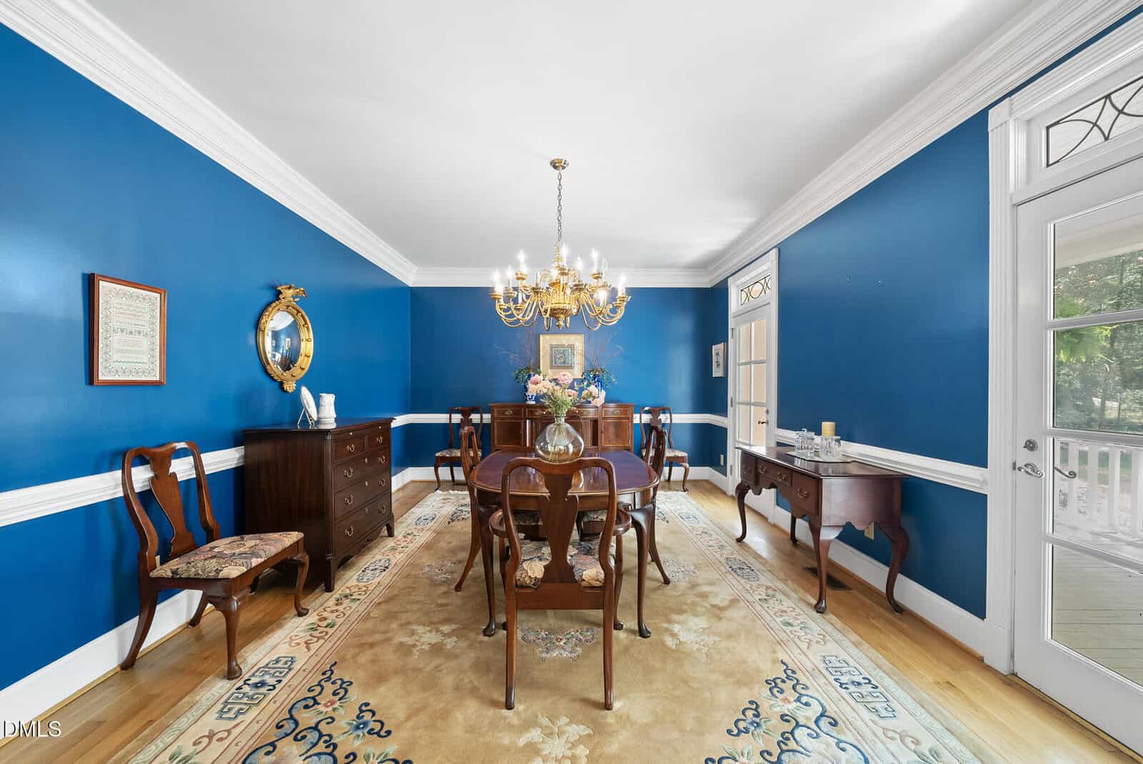 A dining room with blue walls, white trim, a chandelier, wooden table with six chairs, a sideboard, mirror, framed art, and a beige rug on a light wood floor. A glass door leads to an outdoor area.