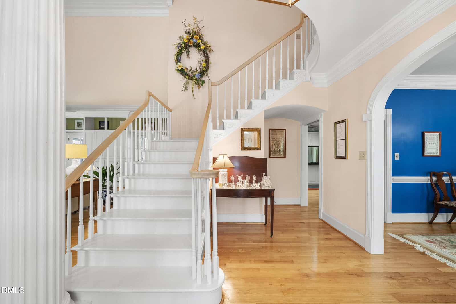 A white staircase with wooden handrails curves upward next to a hallway with light hardwood floors. A table with decorations and a lamp sits below the stairs, and a wreath hangs on the pastel wall above.